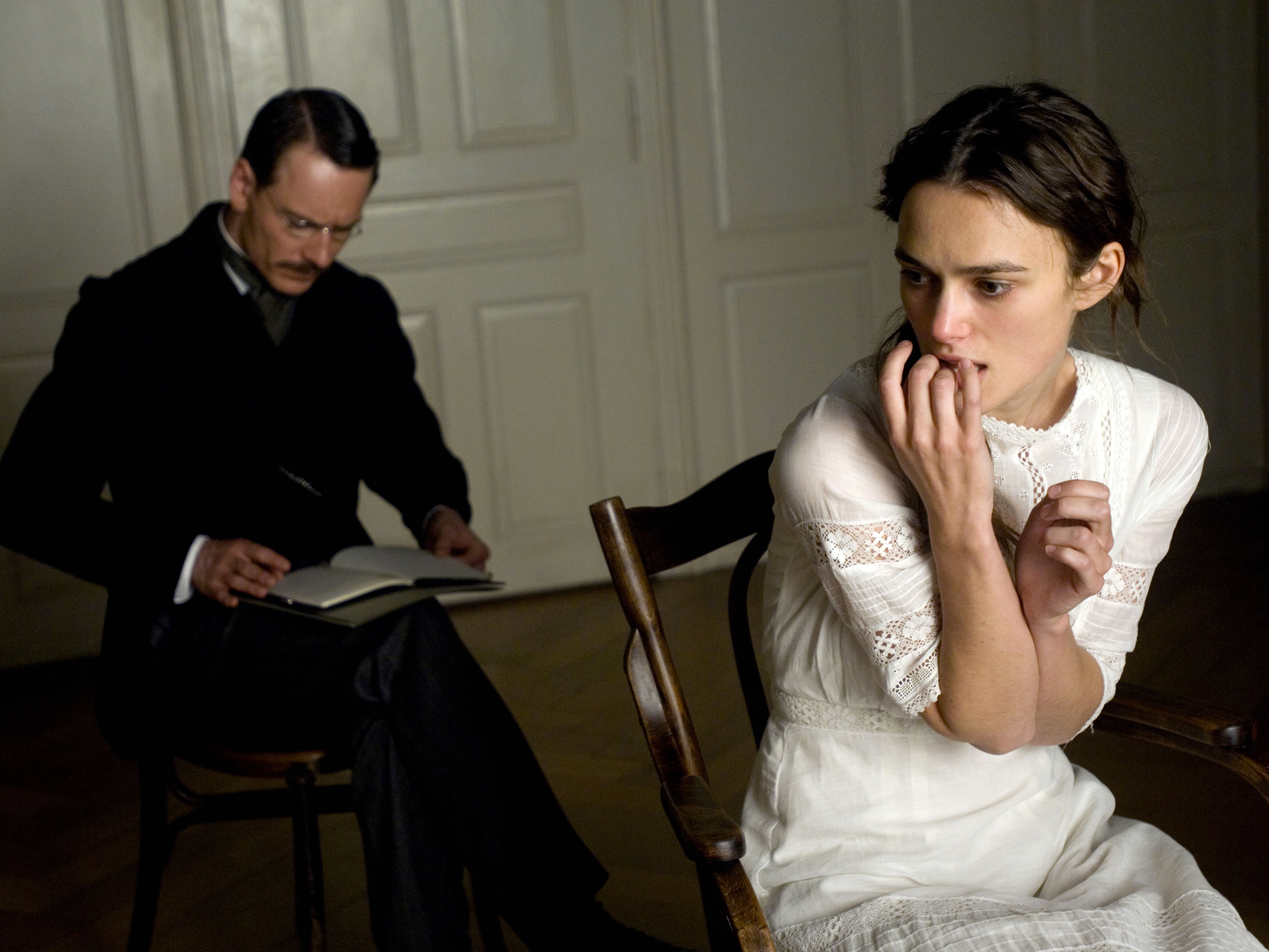 Two figures, a man and a woman, sitting at a table. The man appears to be reading from a book, while the woman is looking pensive. The image has a sombre, serious tone.