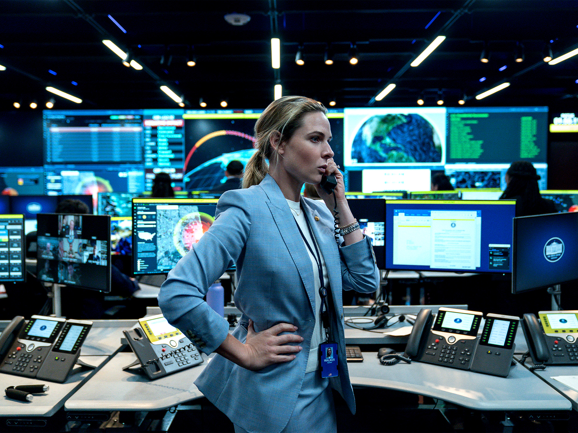 Woman in grey blazer speaking on phone in high-tech control room with multiple monitors displaying maps and data screens.