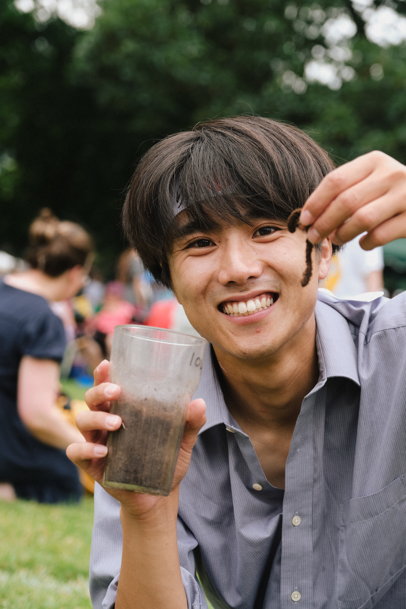 Young Asian man with brown fringe hairstyle smiling whilst holding pint glass outdoors, people and green trees in background.