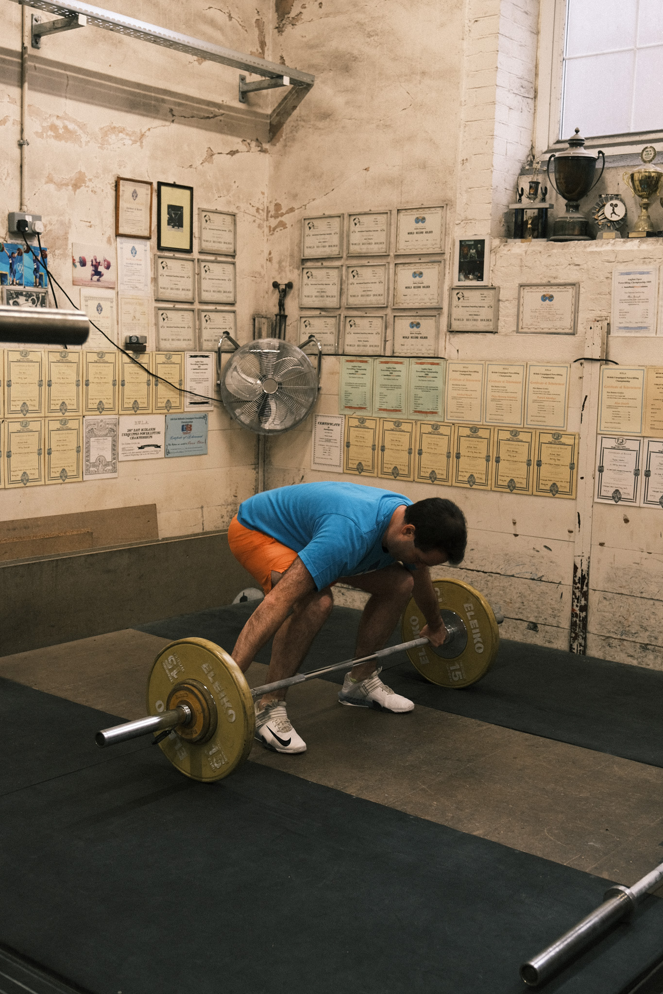 Man in blue shirt and orange shorts lifting barbell in gym with certificates covering weathered white walls and concrete floor.