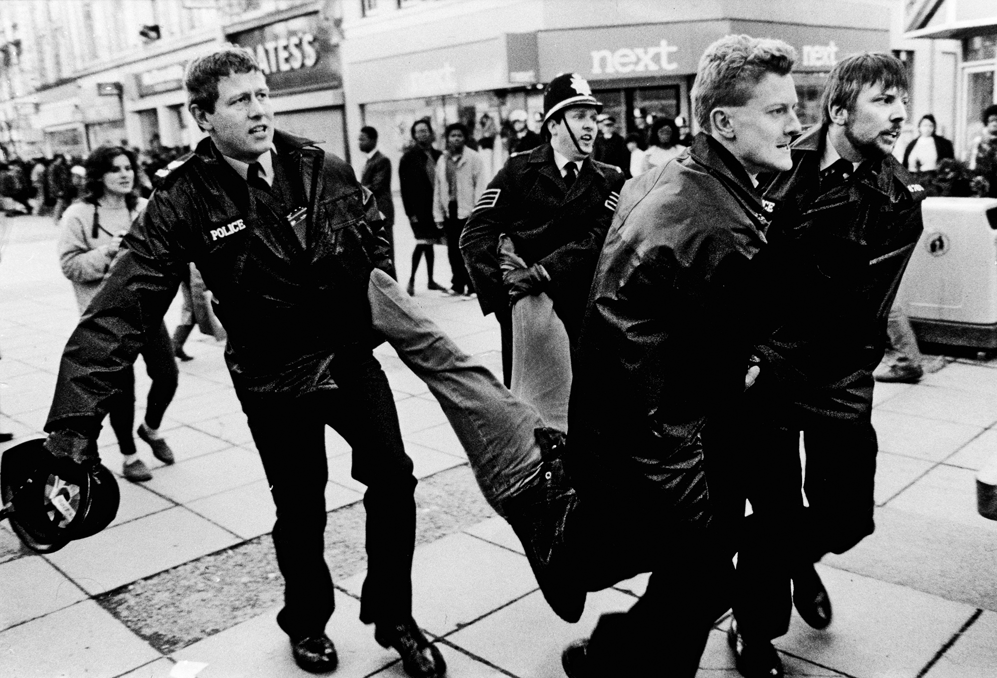 Black and white image showing police officers carrying a person on a street with crowd and shops in background.