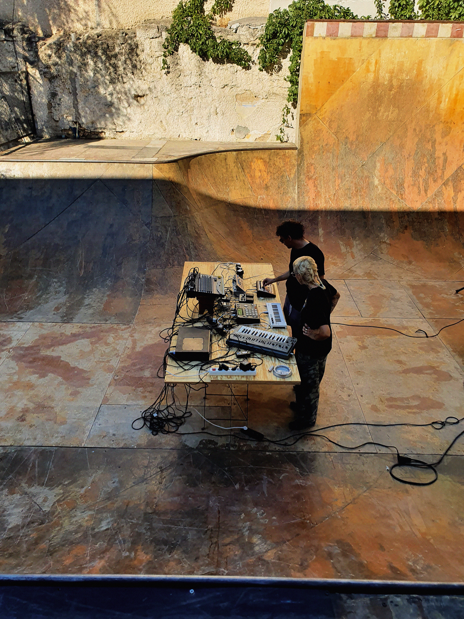 Person playing keyboard at table setup in empty concrete skate bowl with curved orange and grey walls, cables on ground.