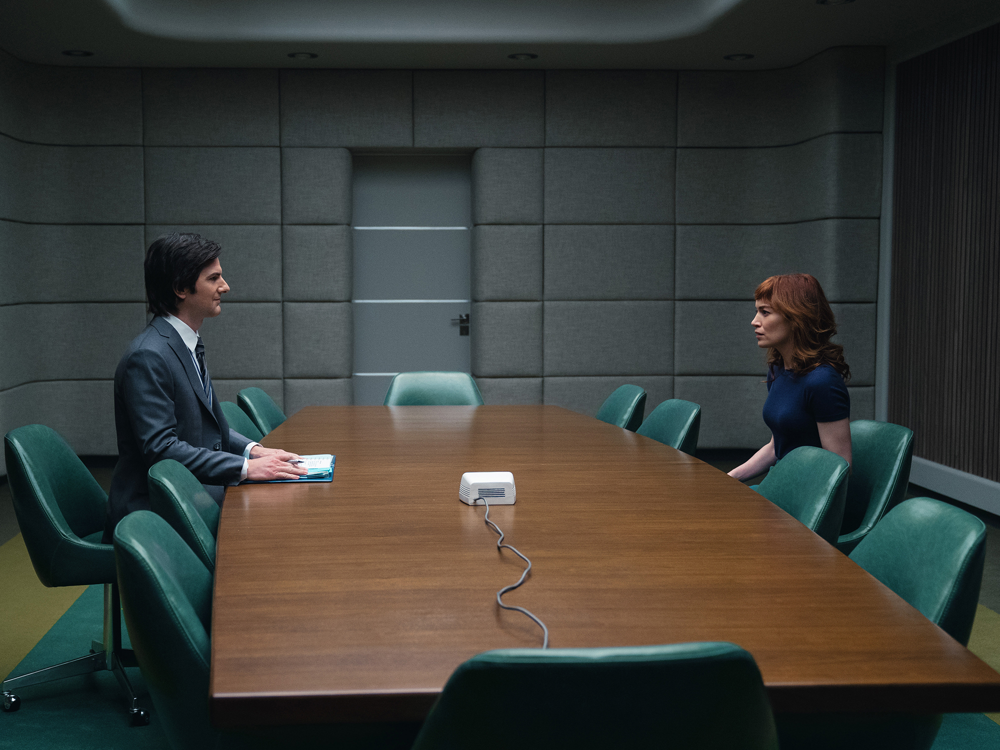 Two people seated at a wooden conference table, one man and one woman, in a room with green panelled walls.