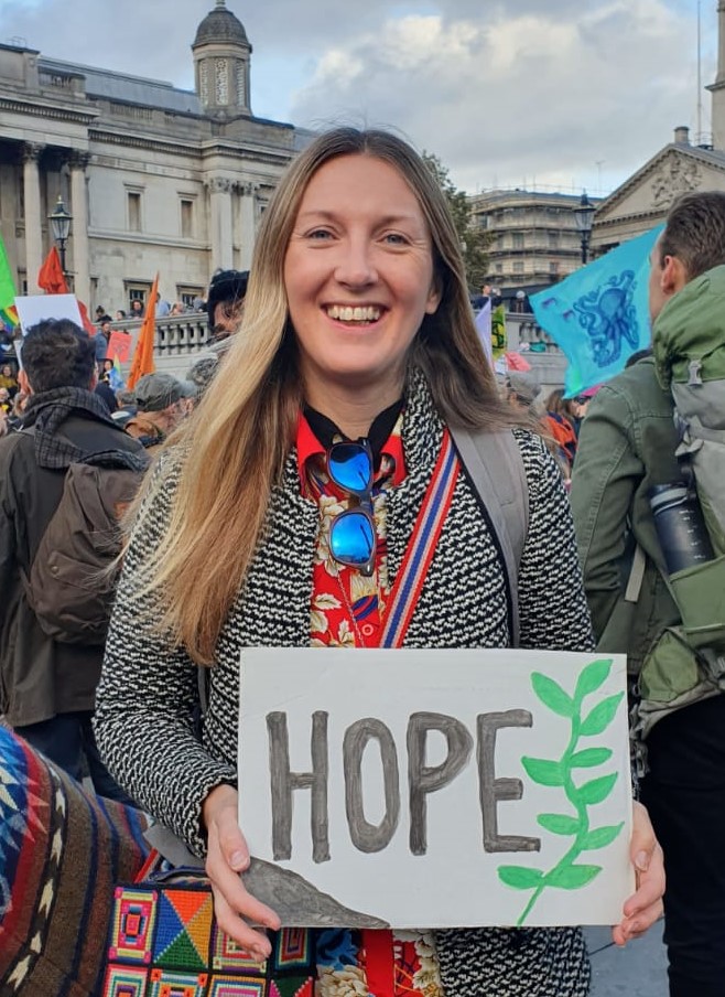 Woman with long blonde hair holding white "HOPE" sign with green plant design at outdoor demonstration in Trafalgar Square, London.