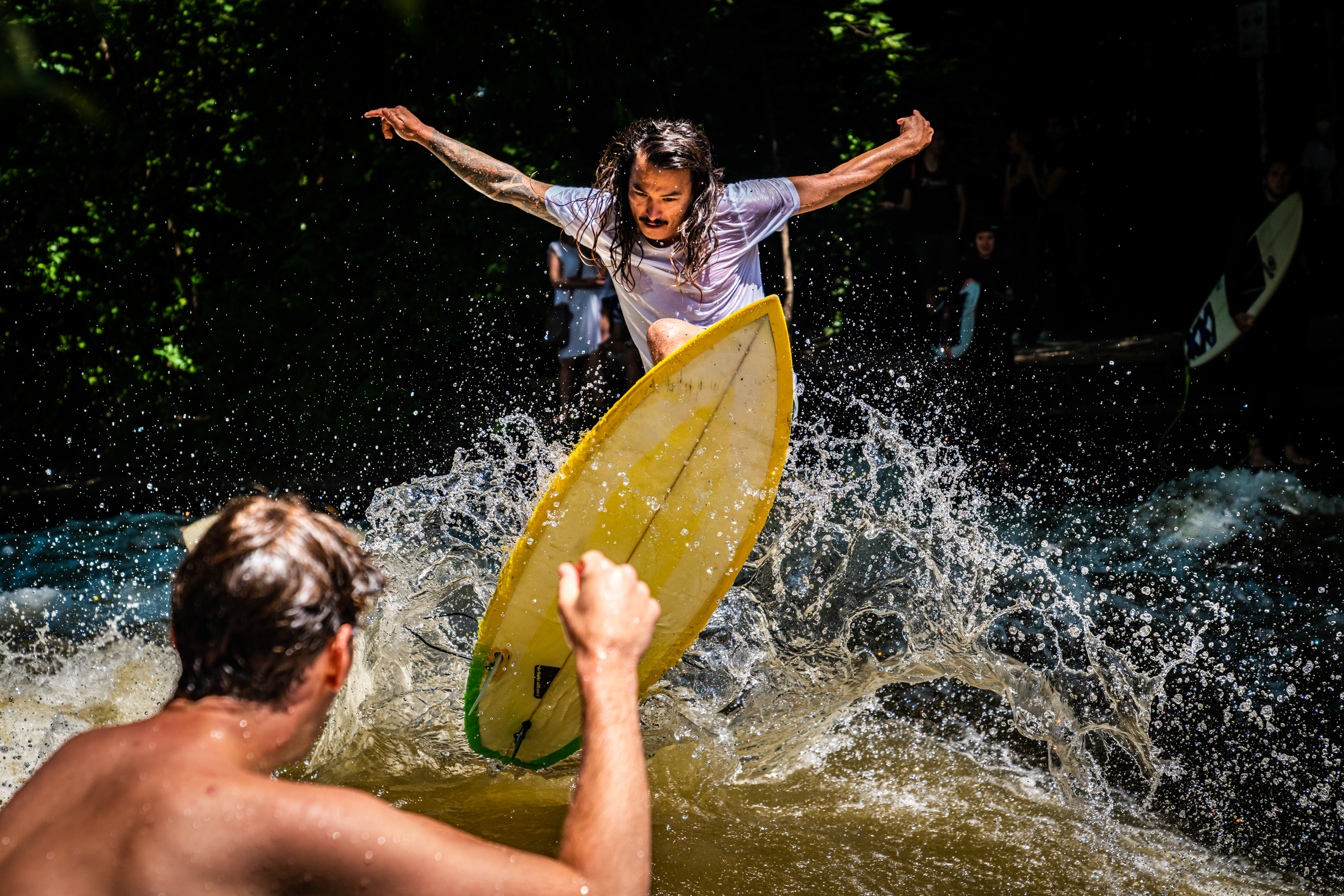 Woman in white top jumping into water with yellow inflatable, arms outstretched. Man's back visible in foreground, water splashing around.