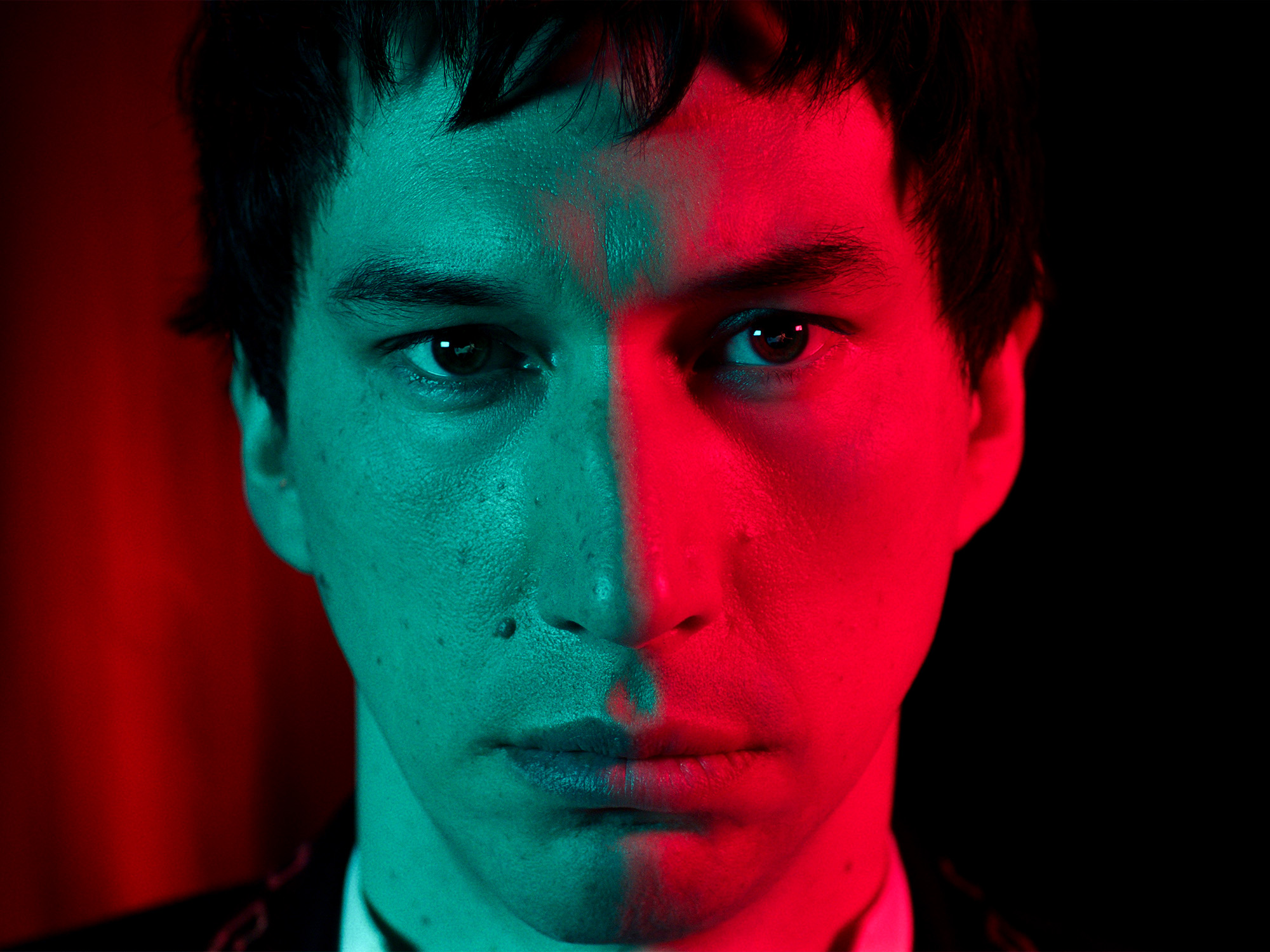 Young man's face lit by contrasting red and cyan lighting, creating a split-colour effect across his features against a dark background.