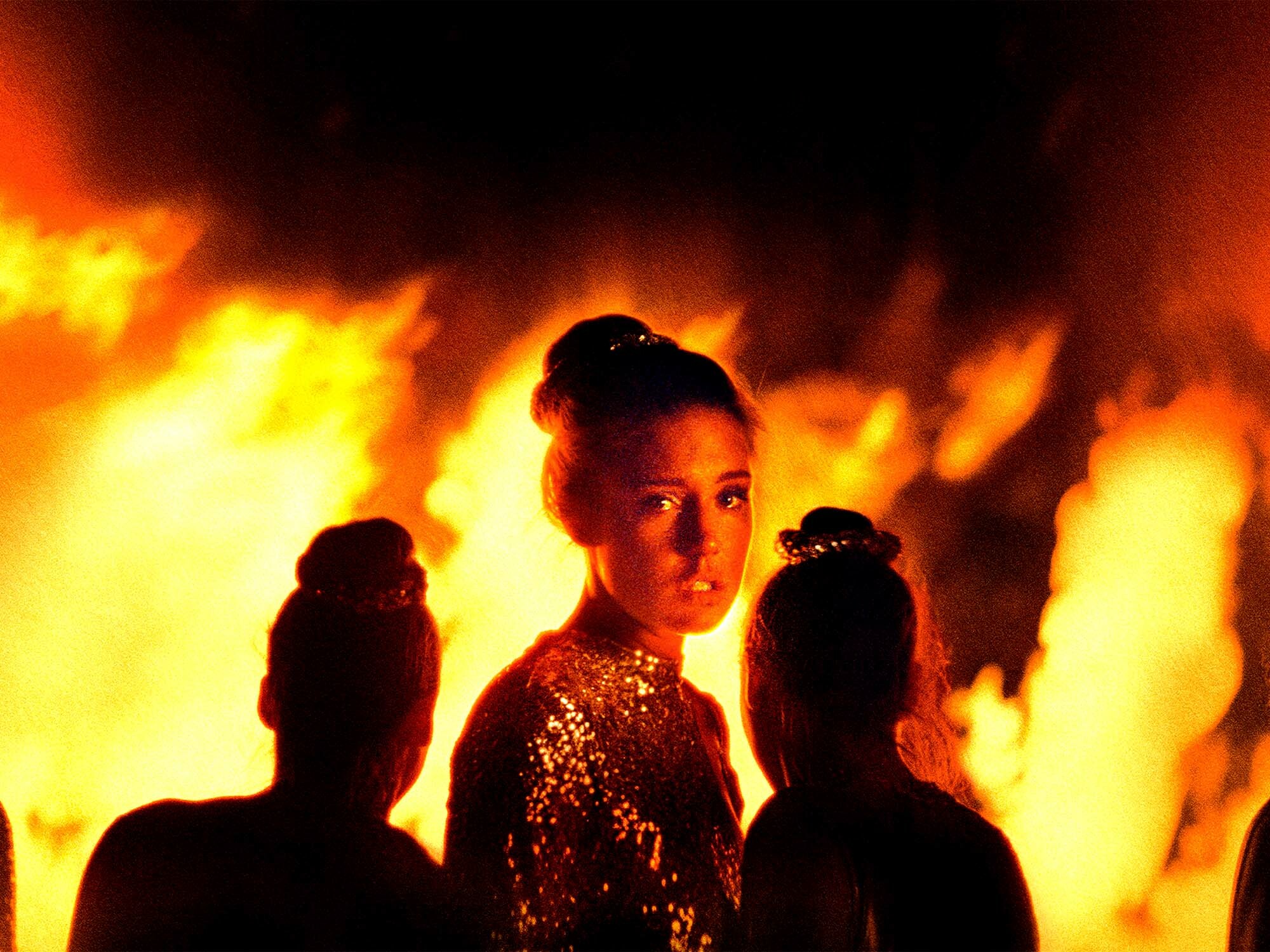 Silhouettes of women in ornate costumes against a fiery orange and yellow backdrop.