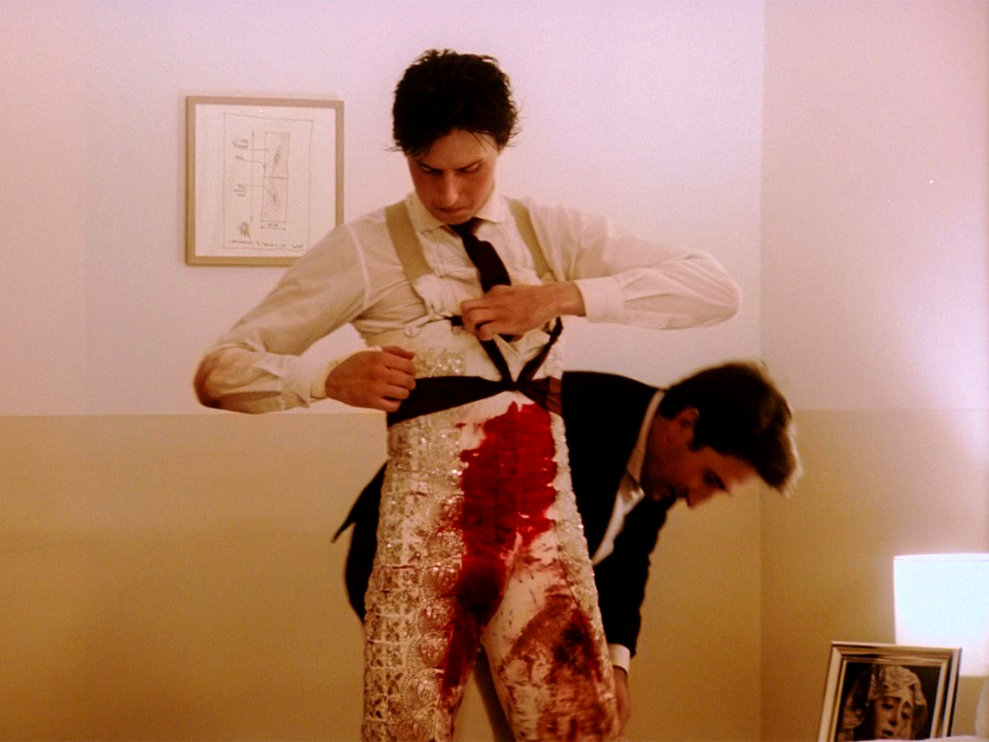 Man in white shirt and braces standing over person in black clothing with red stains, pink and beige interior with framed pictures.