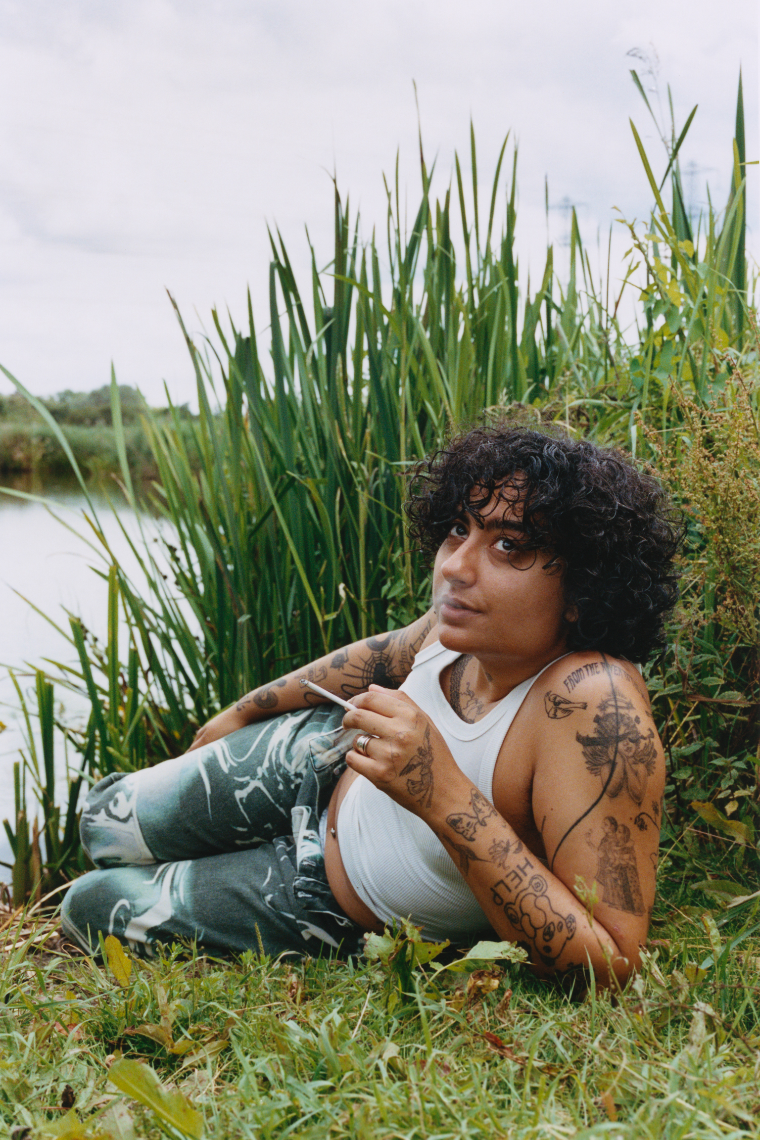 Tattooed person with curly hair in white vest and jeans sits by water's edge amongst tall green reeds under overcast sky.