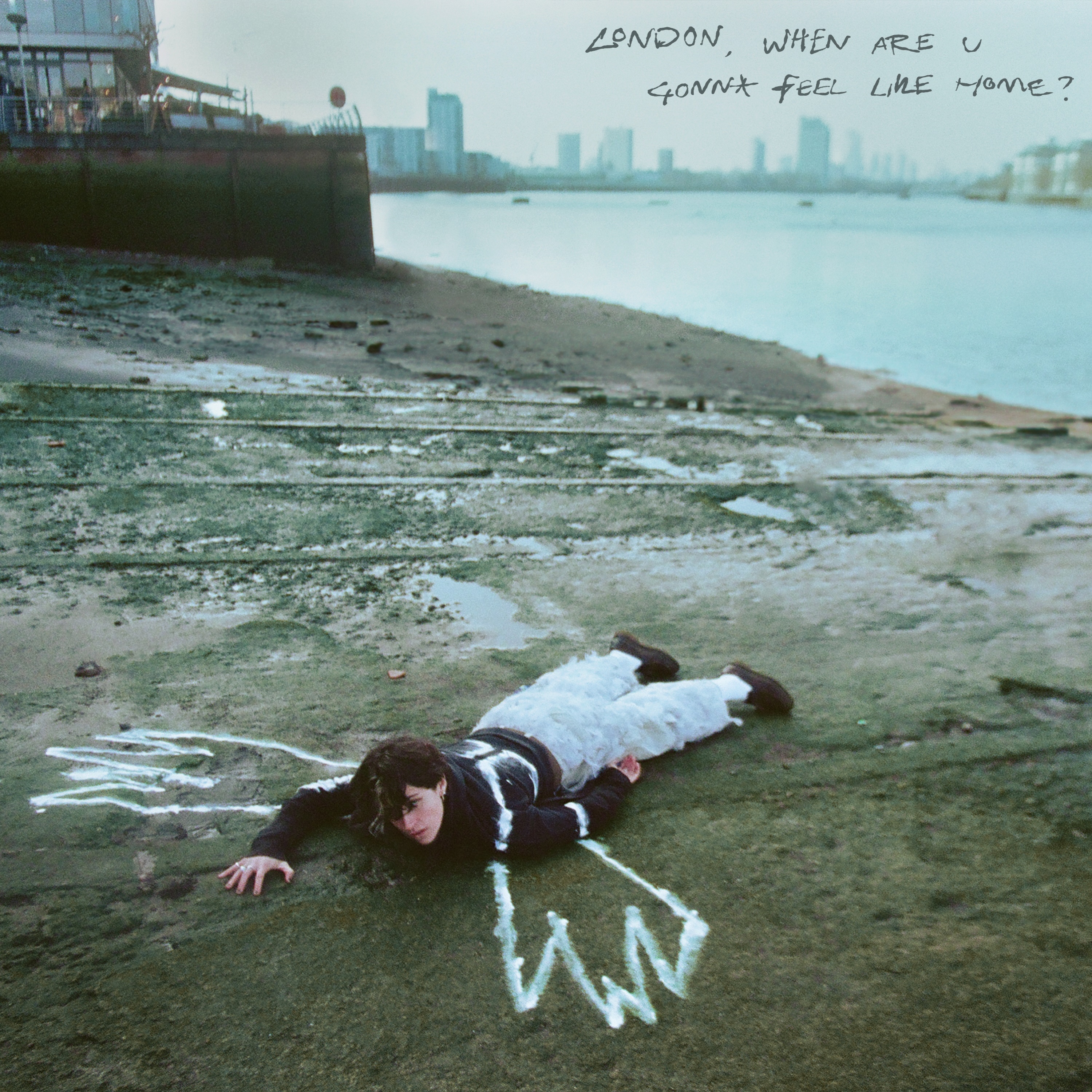 Person lying face-down on muddy Thames foreshore with white chalk outline around body, London skyline visible across grey water.