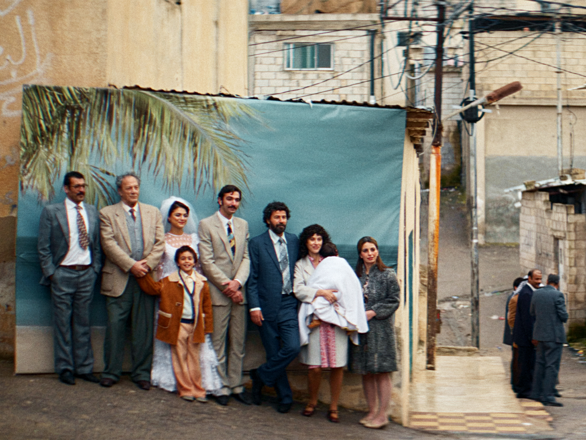 Group of people in formal attire standing against blue wall with palm frond decoration, concrete buildings and power lines visible.
