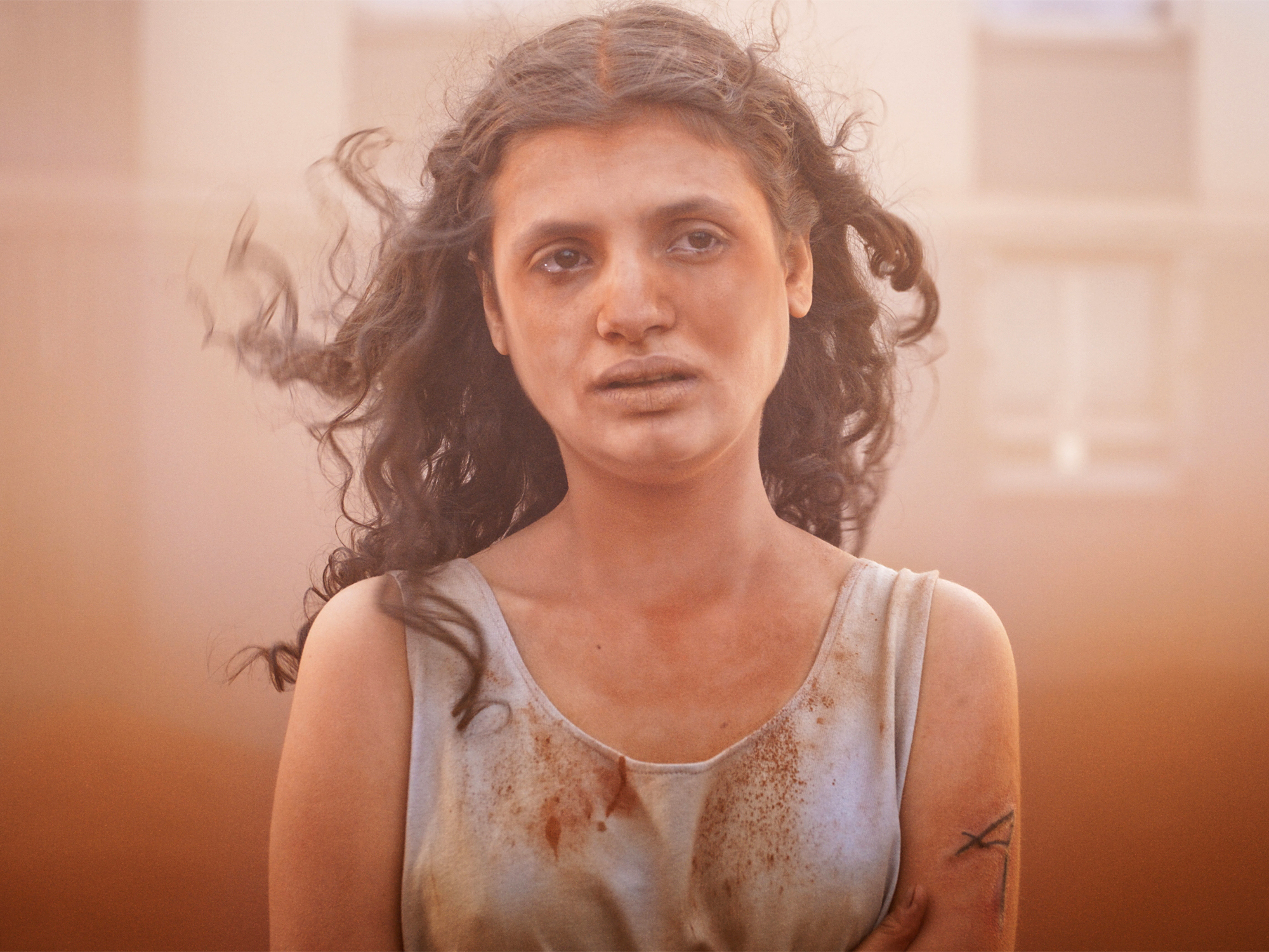 Young woman with curly dark hair wearing a white tank top, with a pensive expression on her face.