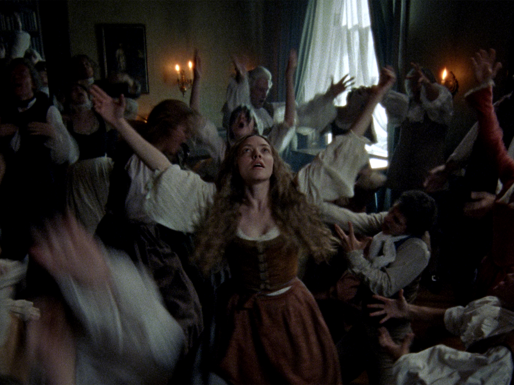 Woman with long brown hair in dark bodice and brown skirt, arms raised, surrounded by crowd in period clothing in dimly lit room.