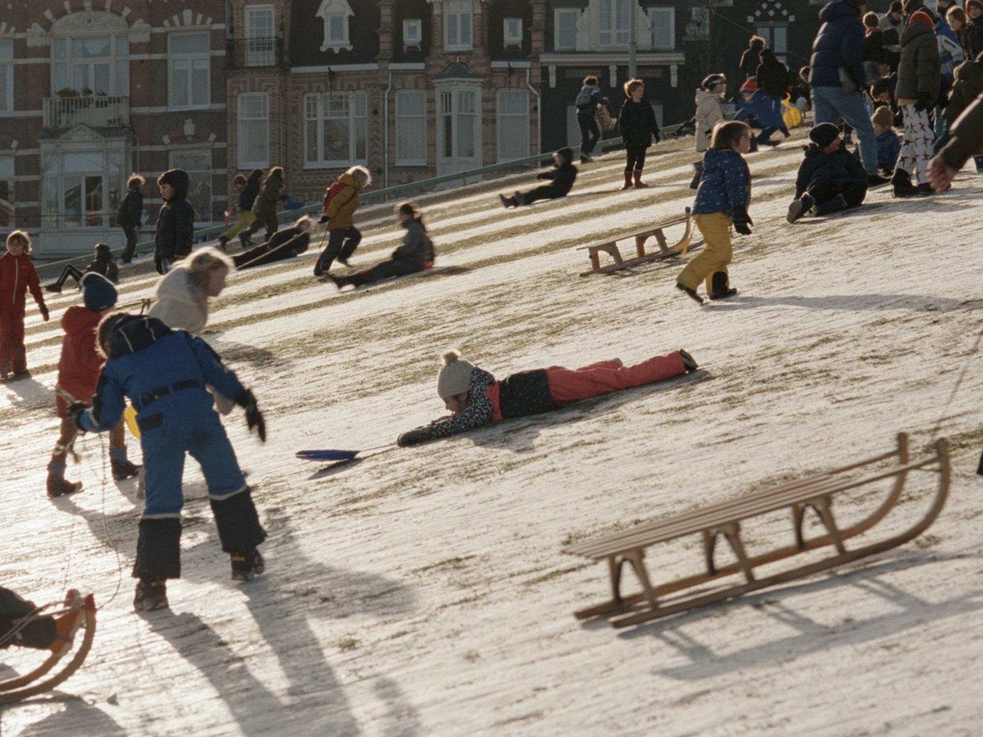 Crowd of people sledging on snowy slope in city, with sleds and buildings in background.