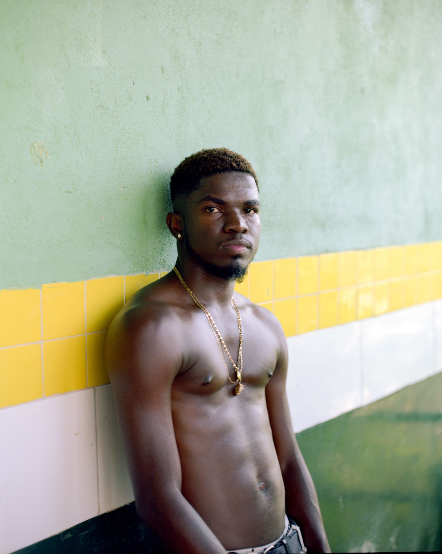 Shirtless Black man wearing gold chain necklace against painted wall with horizontal green, yellow and white stripes.