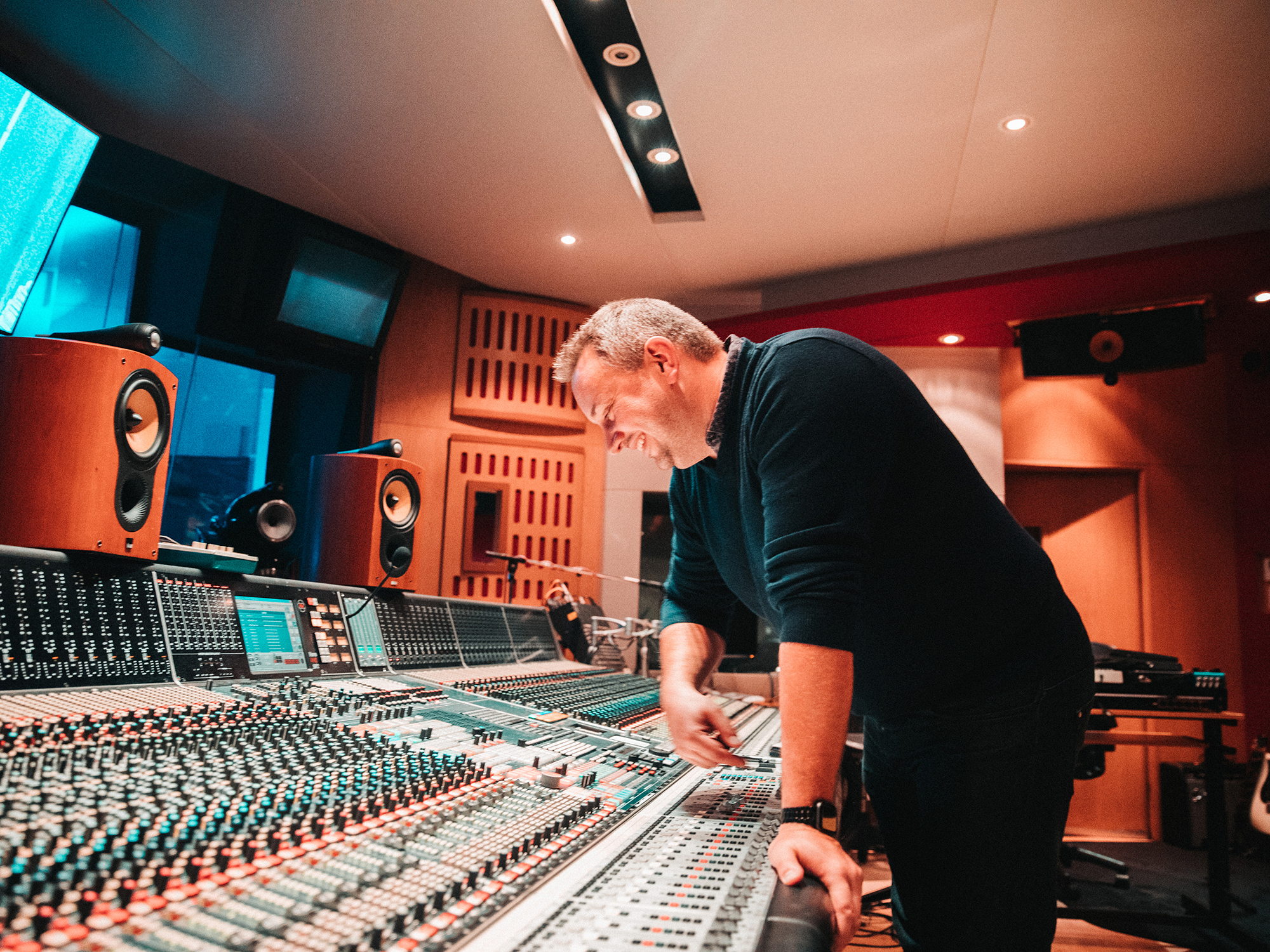 A man, wearing a dark shirt and trousers, working at a professional audio mixing console at Abbey Road Studios. The image shows a variety of audio equipment and speakers.
