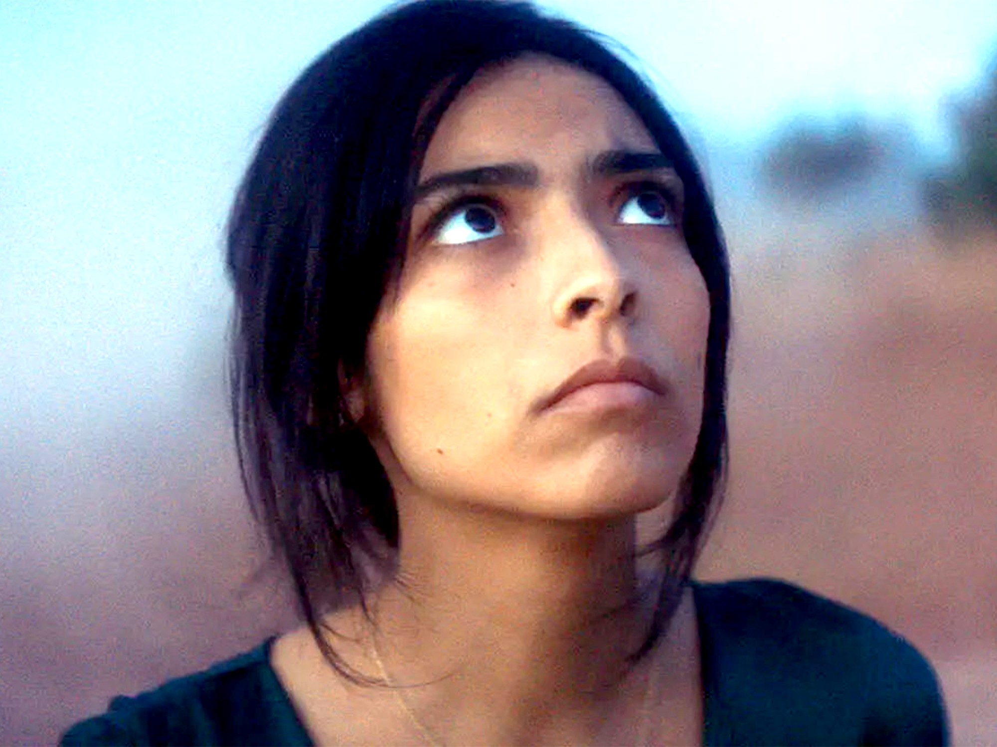Woman with dark shoulder-length hair looking upwards, wearing black top against blurred blue and brown background.