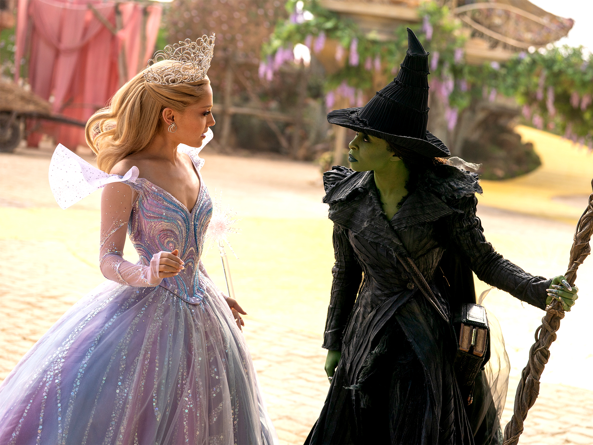 Blonde woman in white dress faces green-skinned figure in black witch hat and robes inside ornate pink-toned interior with arched architecture.