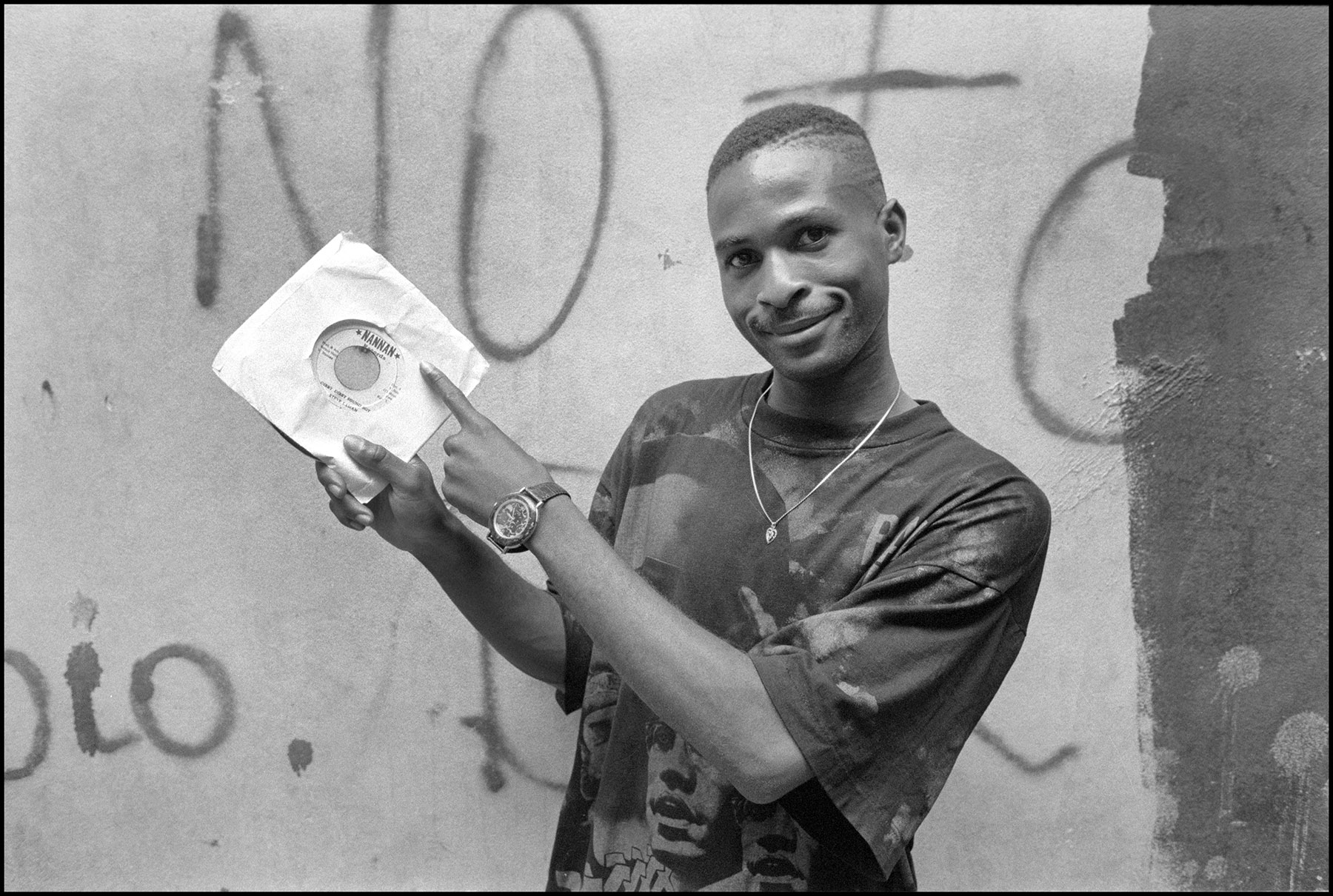 Black and white image: smiling Black man holding white cassette tape player against graffitied wall background.