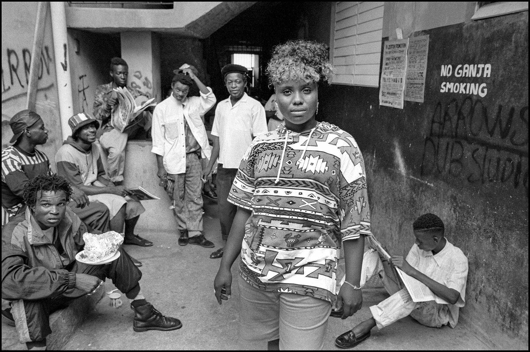Black and white image showing woman in patterned top walking past seated and standing people outside building with "NO GANJA SMOKING" sign.