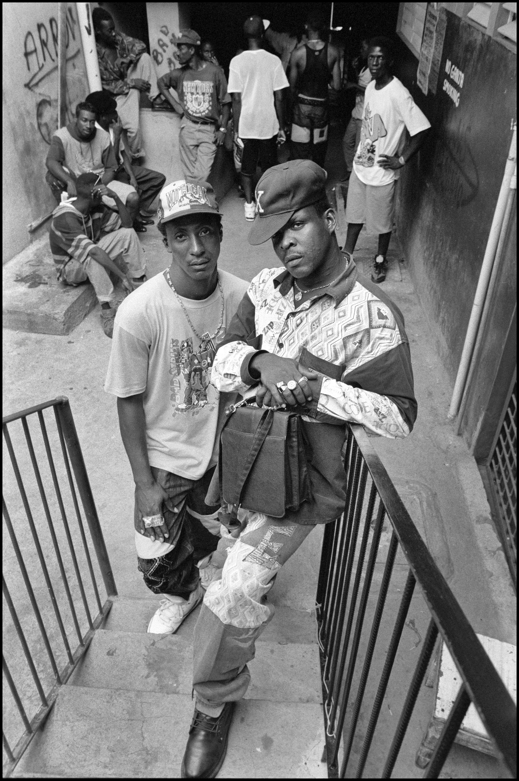 Black and white image showing group of young men on concrete steps and landing, two in foreground wearing caps and casual clothing.