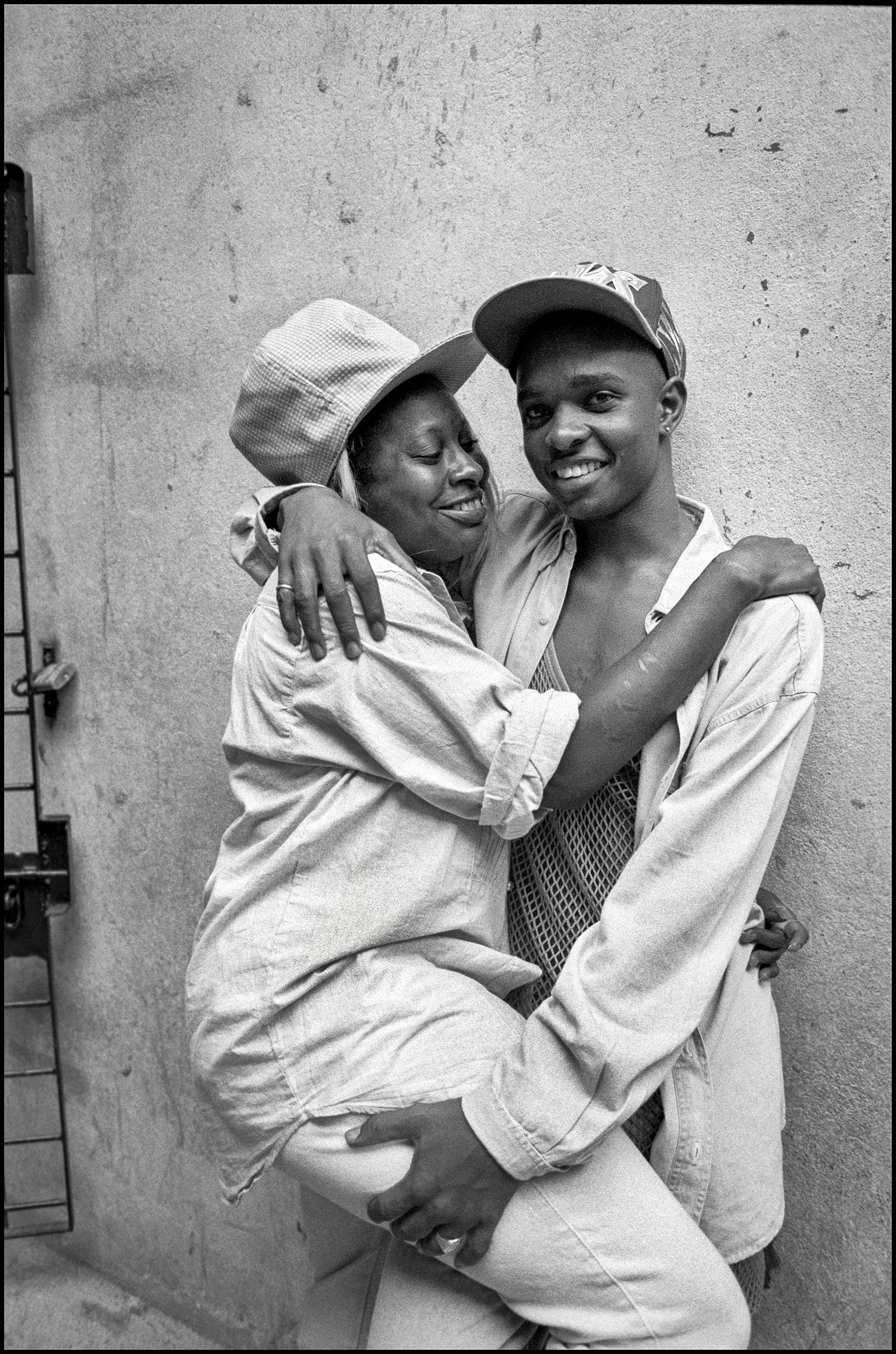 Two young women embracing and smiling, wearing caps and loose clothing, against weathered wall. Black and white photograph.