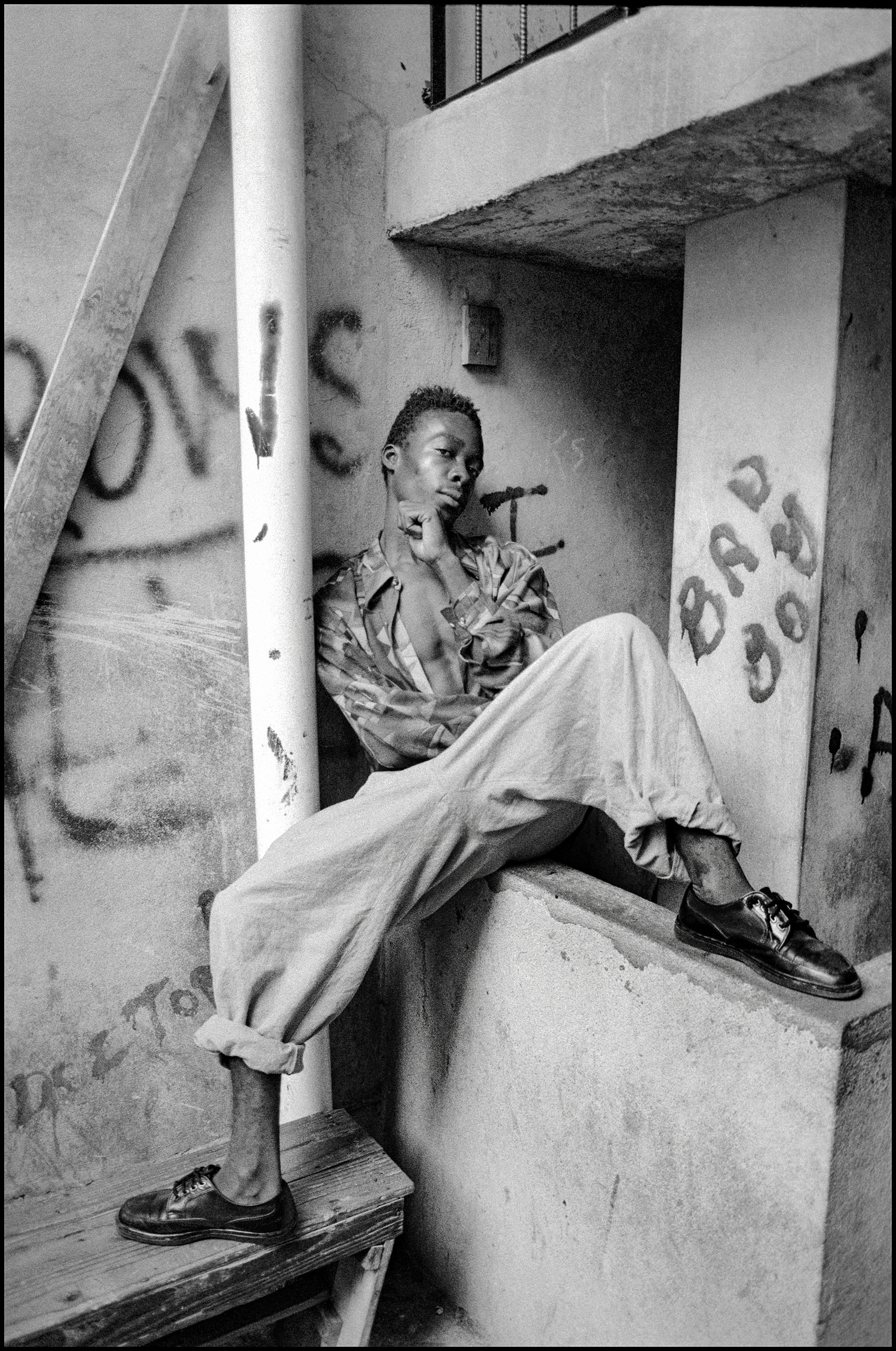 Black and white image of person sitting on concrete steps wearing patterned shirt and wide trousers, graffitied walls visible.