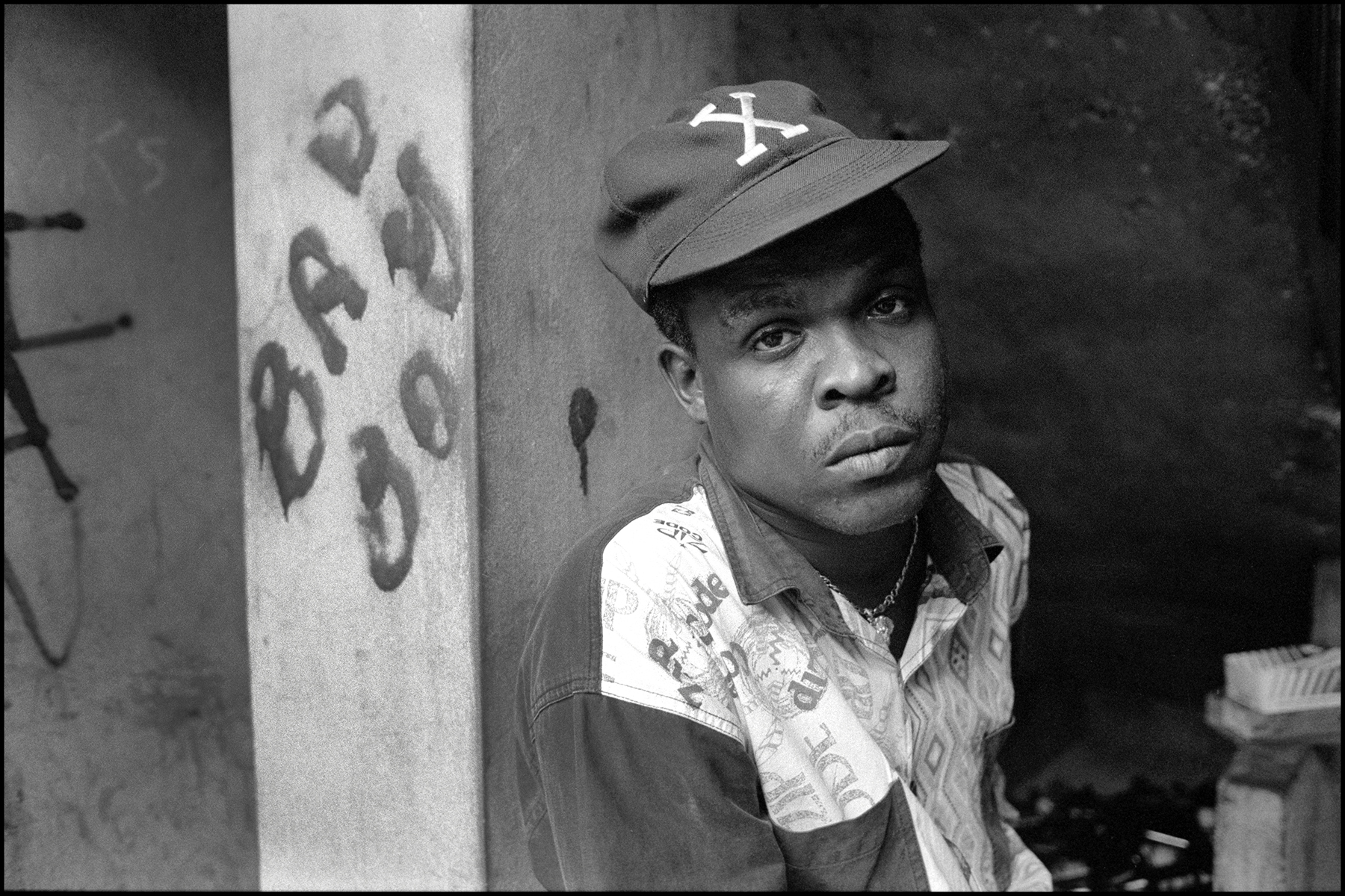 Black and white portrait of young Black man wearing baseball cap with "A" logo and patterned shirt, standing beside white pillar with numbers.