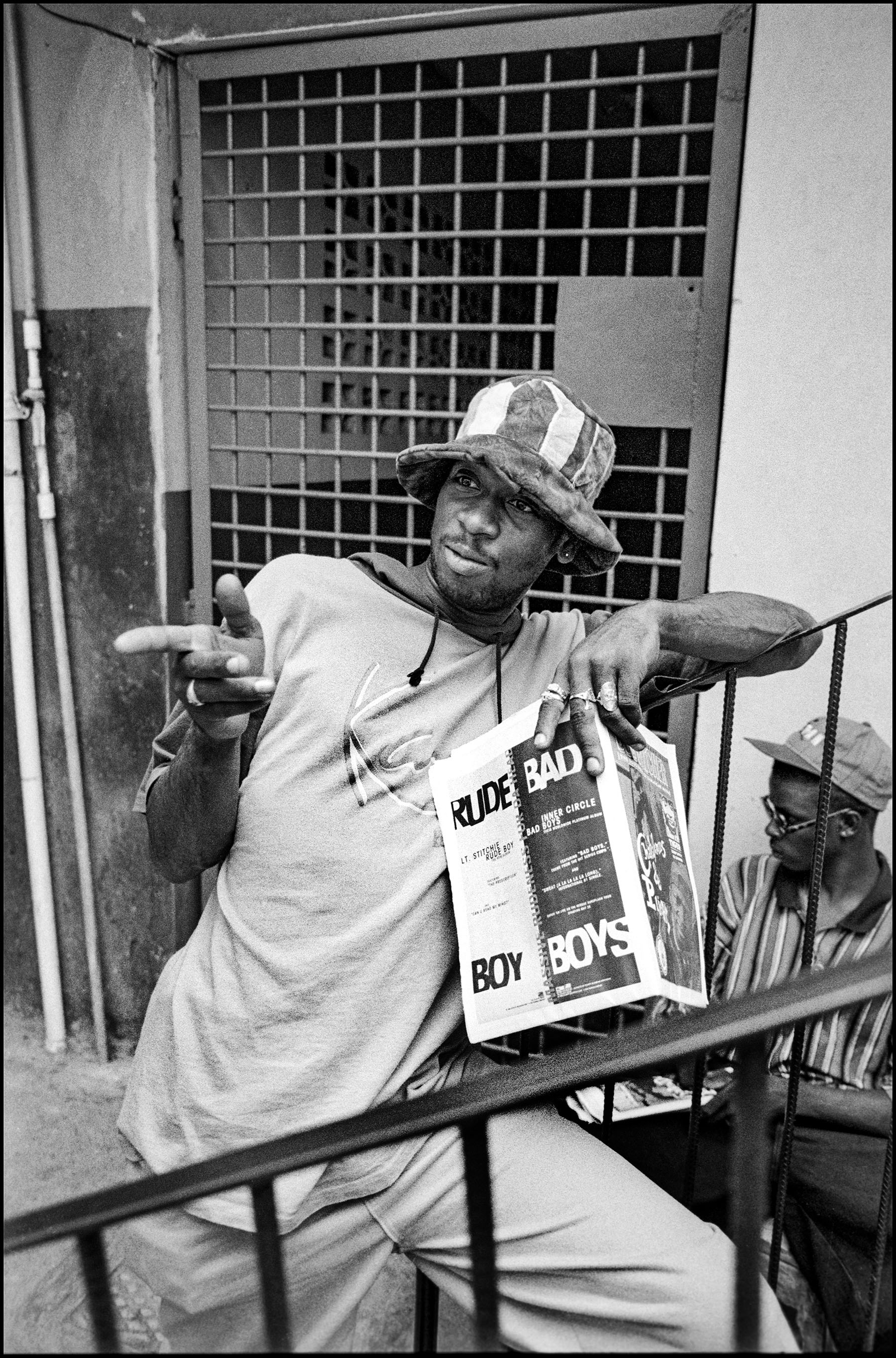 Black and white image of man in bucket hat and light shirt holding magazine, sitting on metal railings with barred window behind.