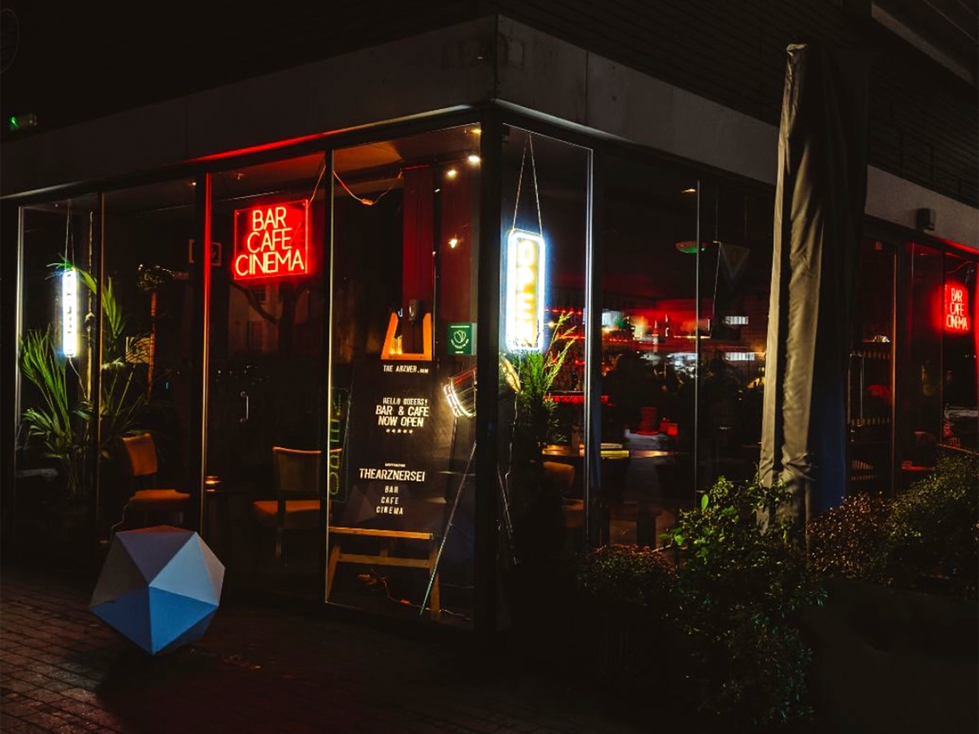 Night-time storefront with large glass windows, red neon "BAR CAFE CINEMA" sign, warm interior lighting, and blue geometric object outside.