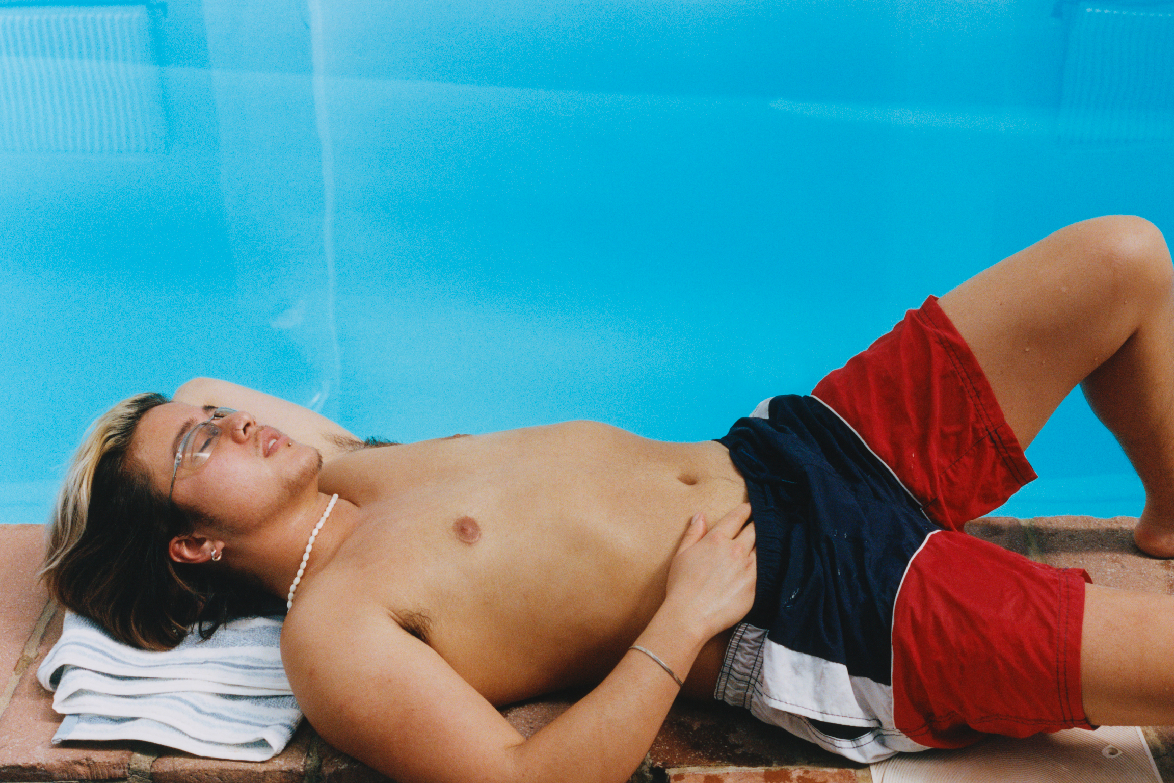 Shirtless man in red and black shorts lying on white towel beside bright blue swimming pool.