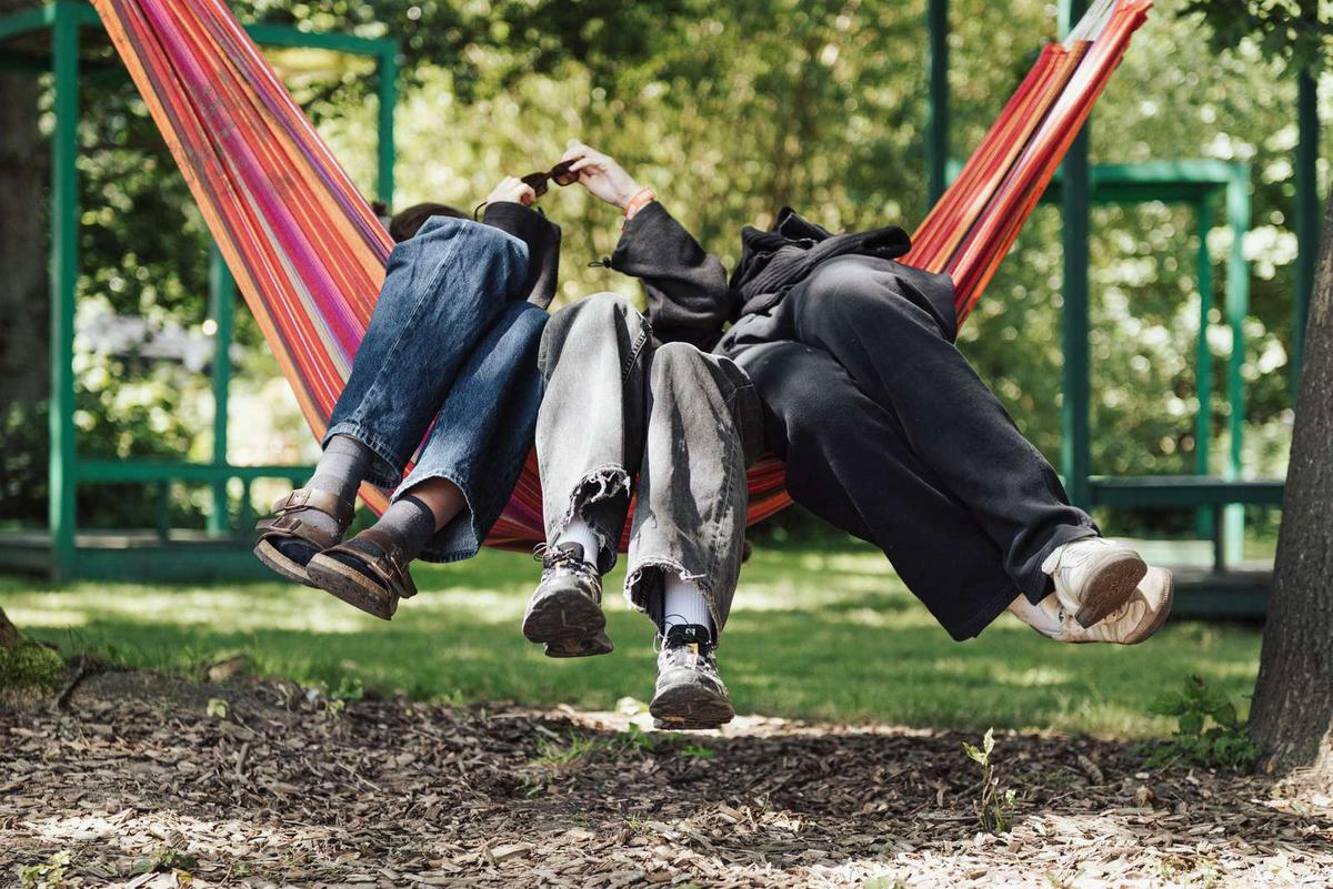 Three people lying in colourful striped hammock in park, legs dangling over sides, wearing jeans and casual trousers.