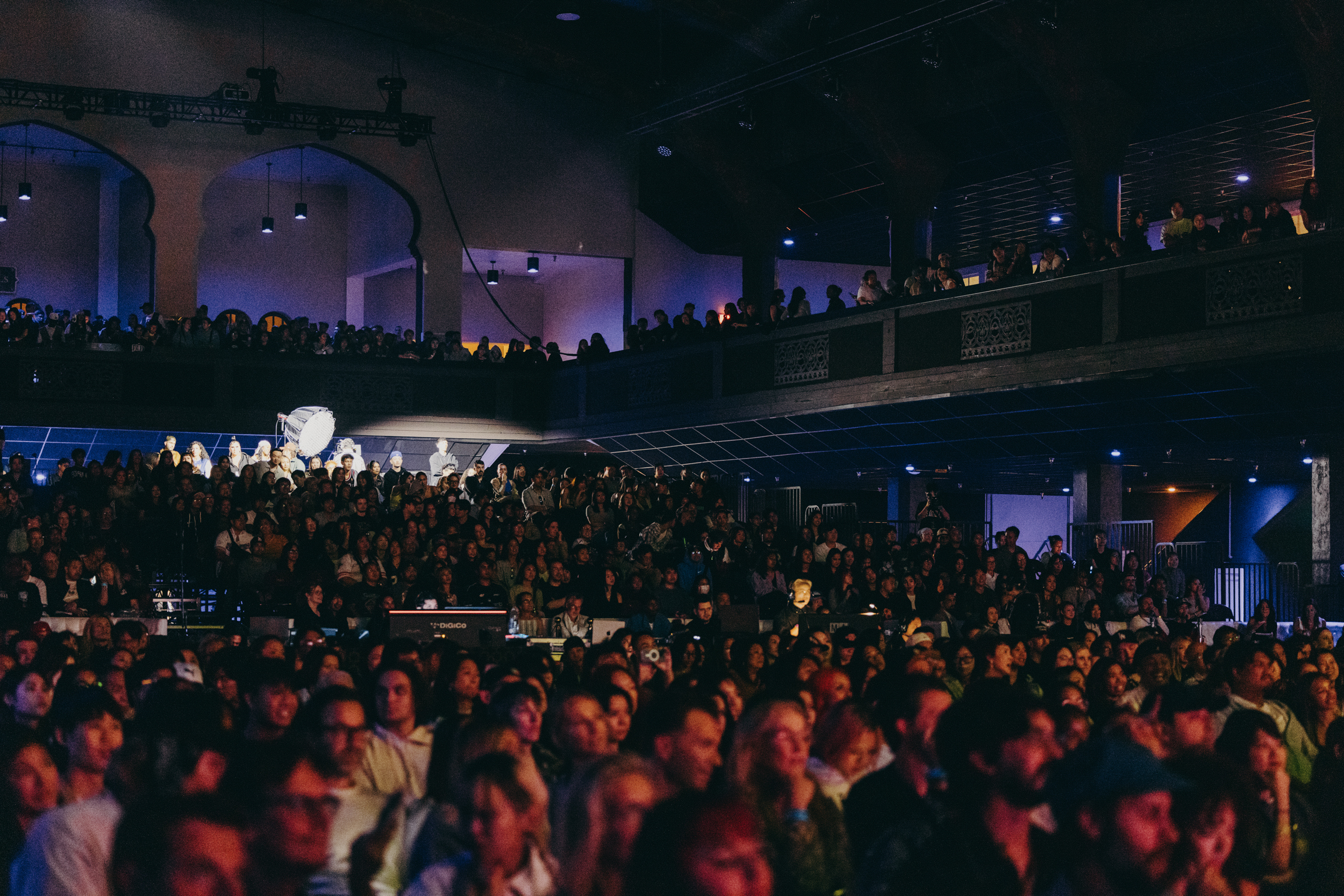 Large indoor venue packed with crowds on multiple levels, dimly lit with purple and blue stage lighting, balcony seating visible above.