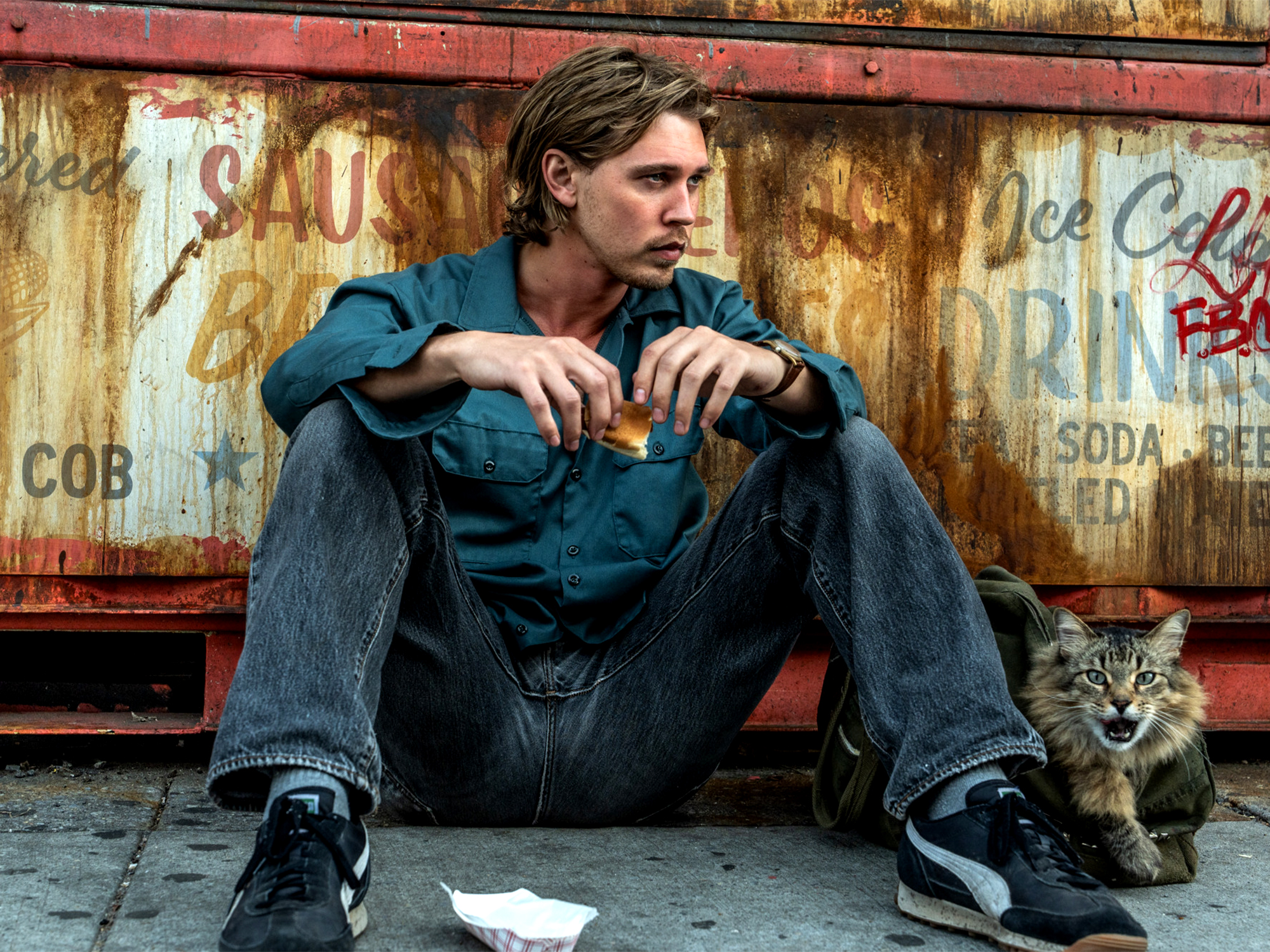 Young man in teal hoodie and jeans sits on ground against weathered metal wall with vintage signage, small tabby kitten beside him.