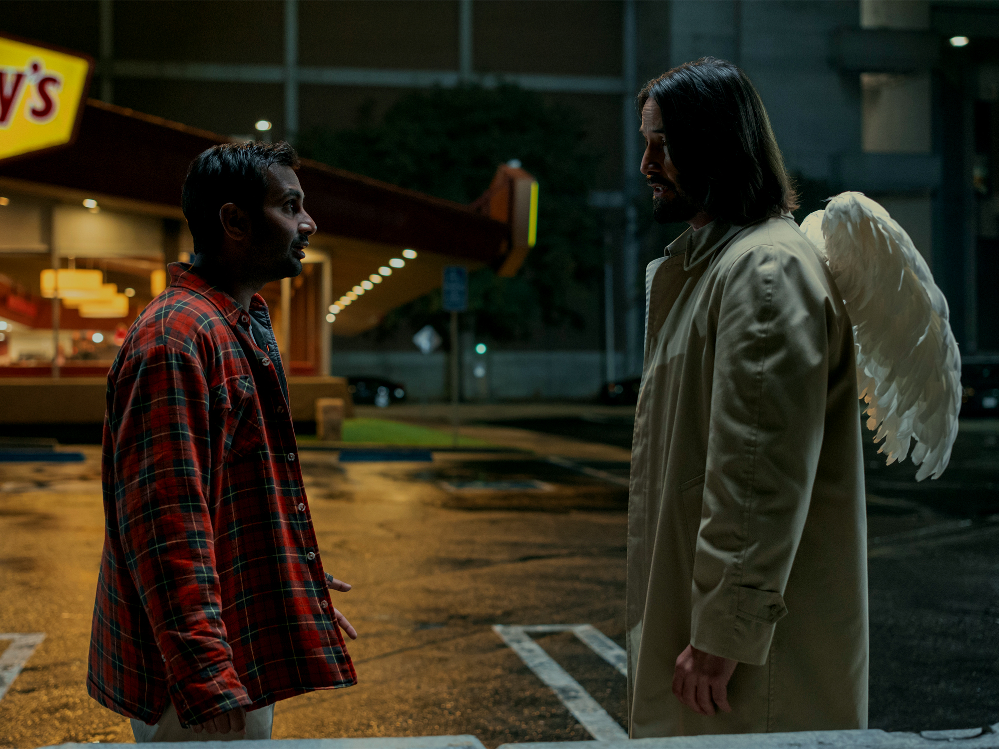 Two men talking outside fast food restaurant at night. One wears red plaid shirt, other in beige coat with white wings on back.