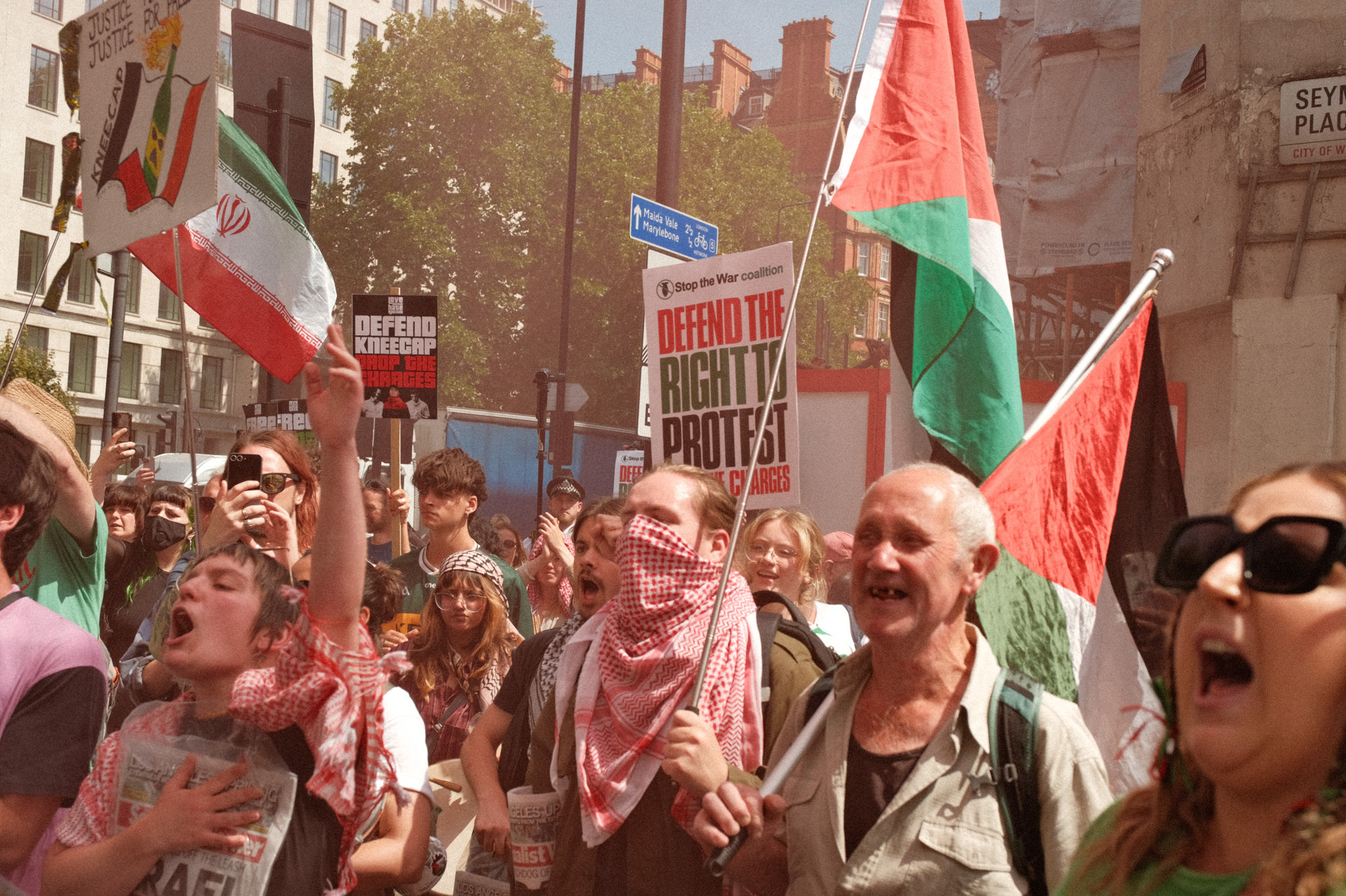 Crowd of protesters holding Palestinian and Iranian flags, carrying signs that read "Defend the right to protest".