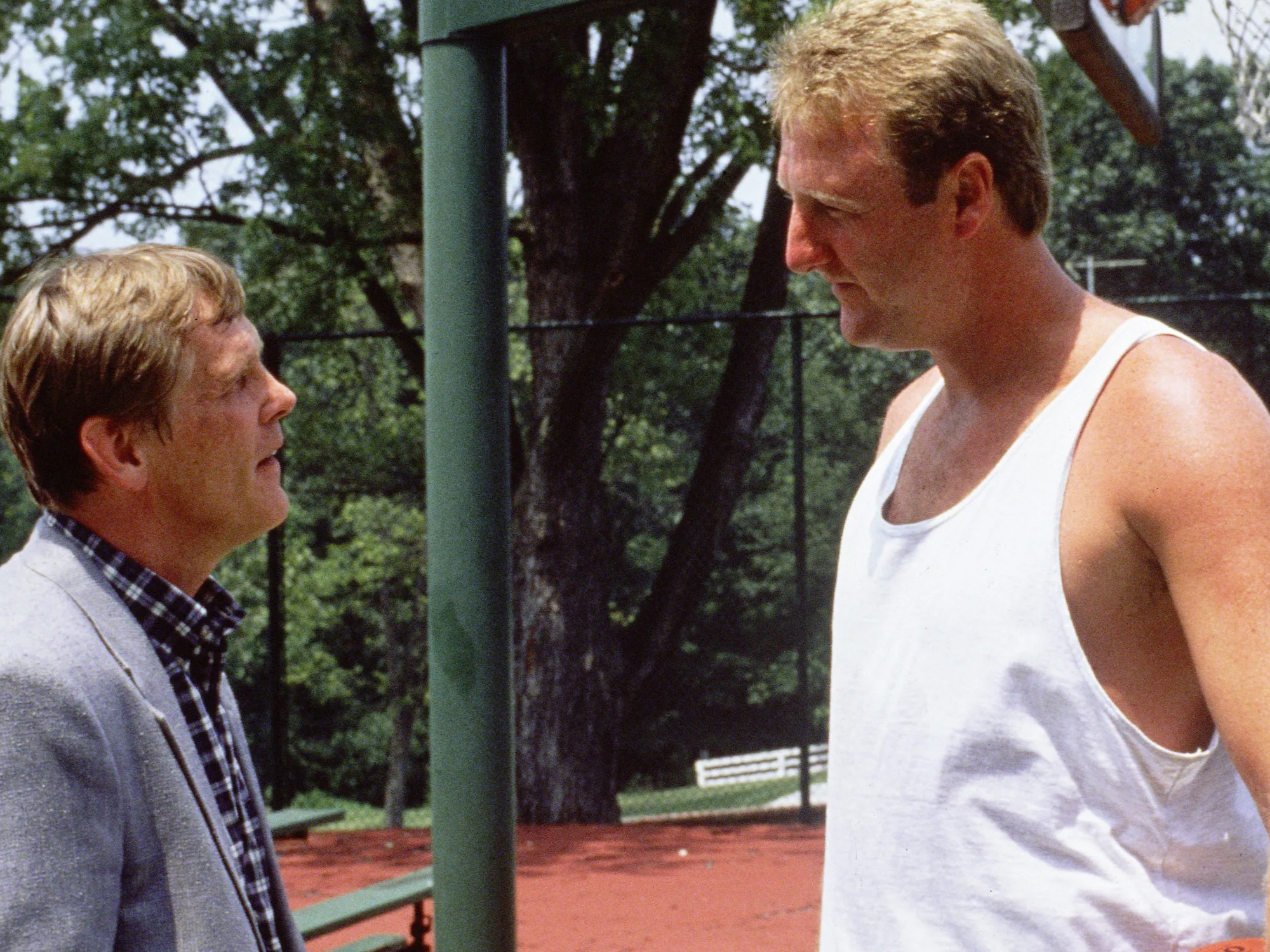 Two men taking on a basketball court, surrounded by trees.
