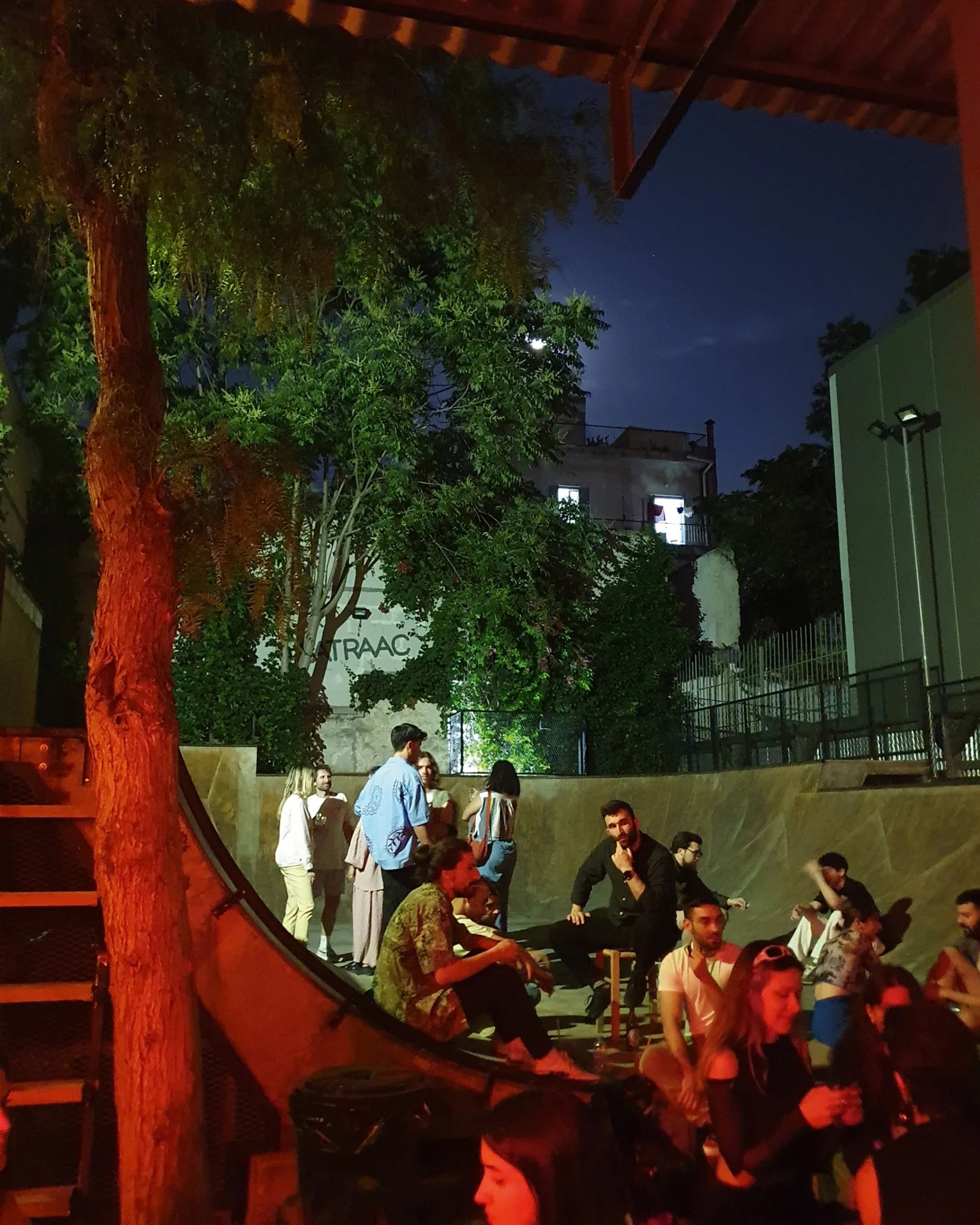 Group of people gathered on outdoor steps at night under string lights, with green trees and buildings in background under dark blue sky.