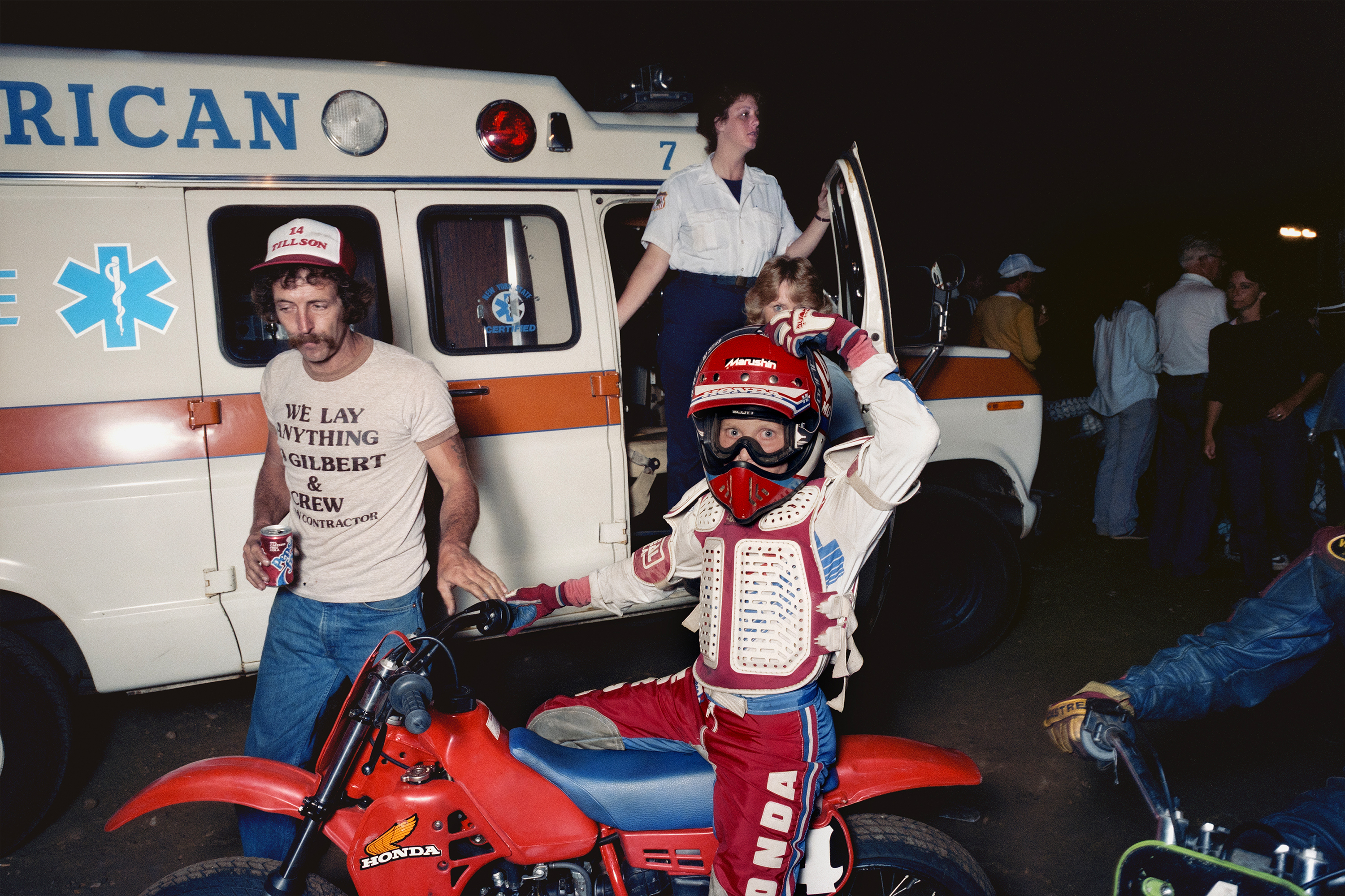 Motocross rider in red gear beside Honda dirt bike with ambulance and medical personnel in background at night event.