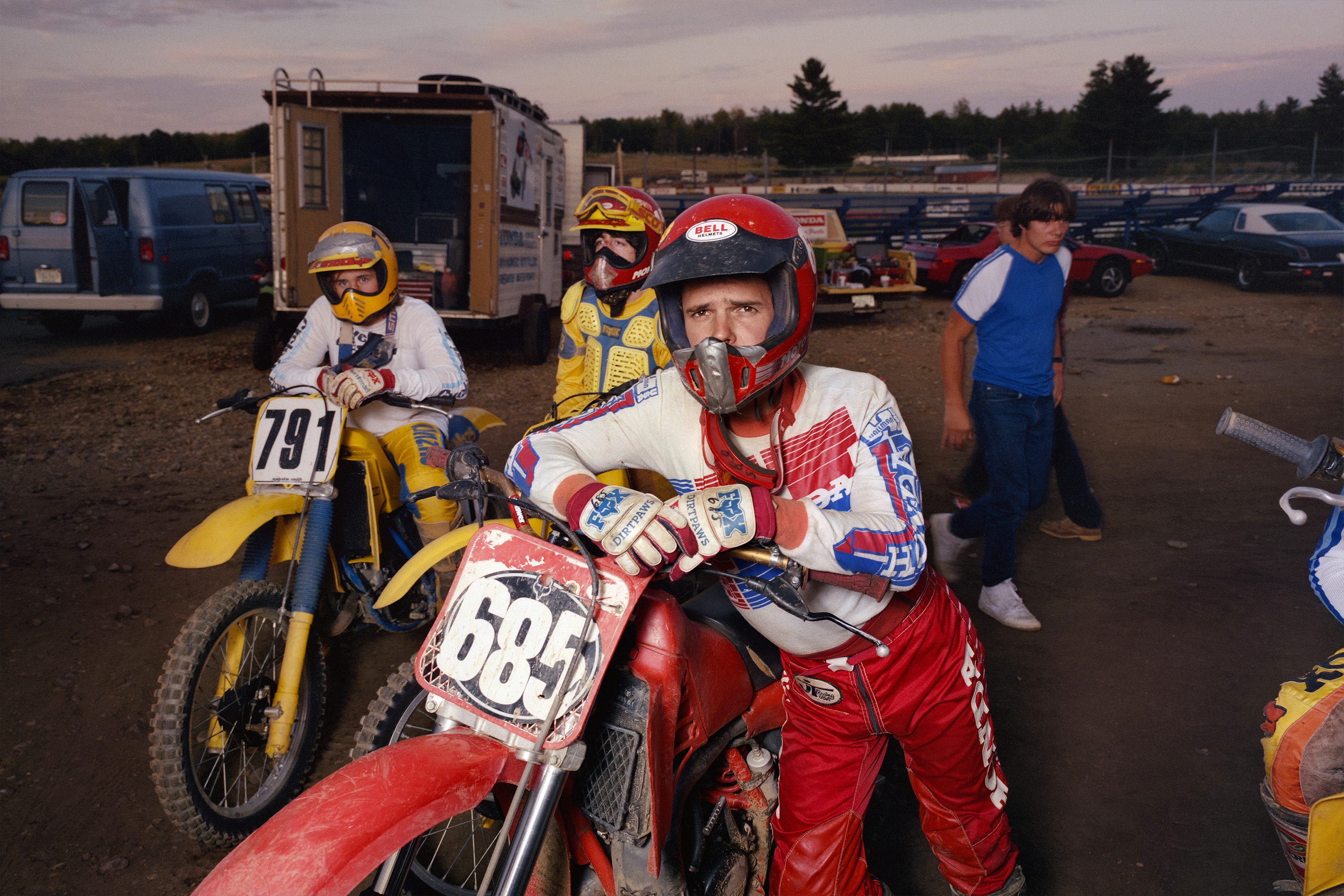 Motocross riders in helmets and racing gear sit on dirt bikes numbered 791 and 685 in crowded paddock area with lorries and cars.