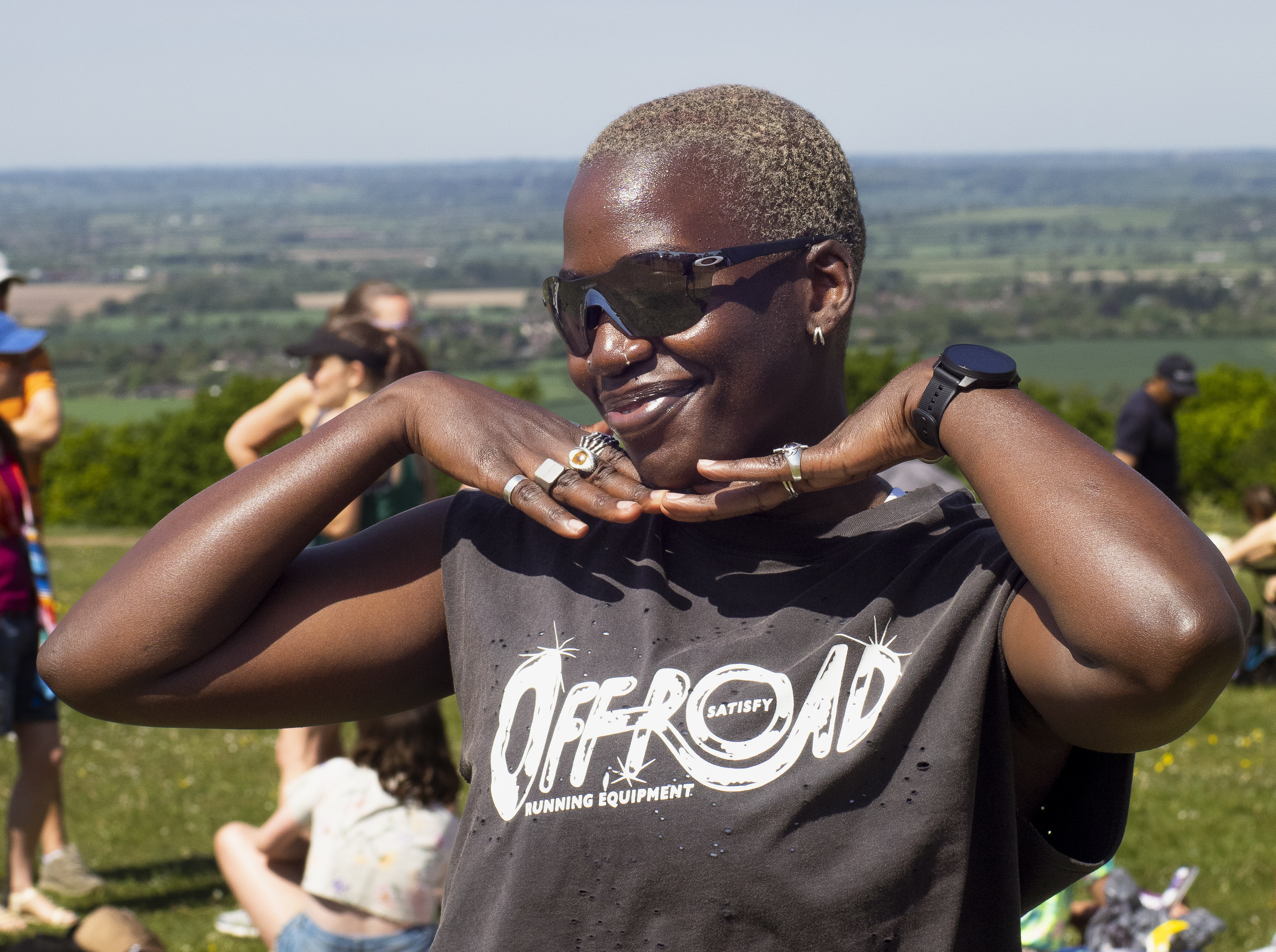 Black woman with short blonde hair and sunglasses smiling at camera, wearing grey "Off Road" t-shirt, countryside landscape behind.