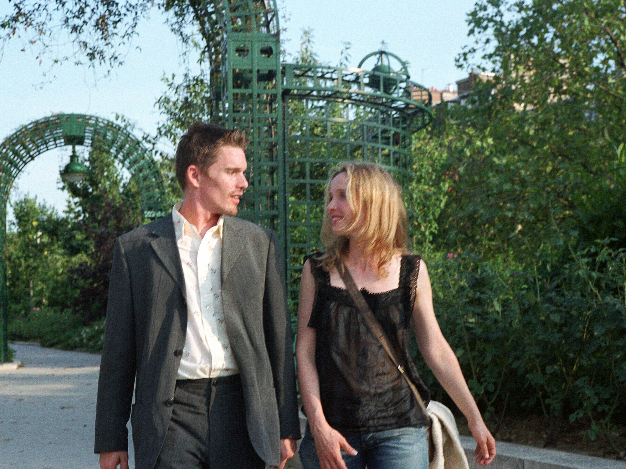 Two people walking together in a park with a green metal arch structure in the background.