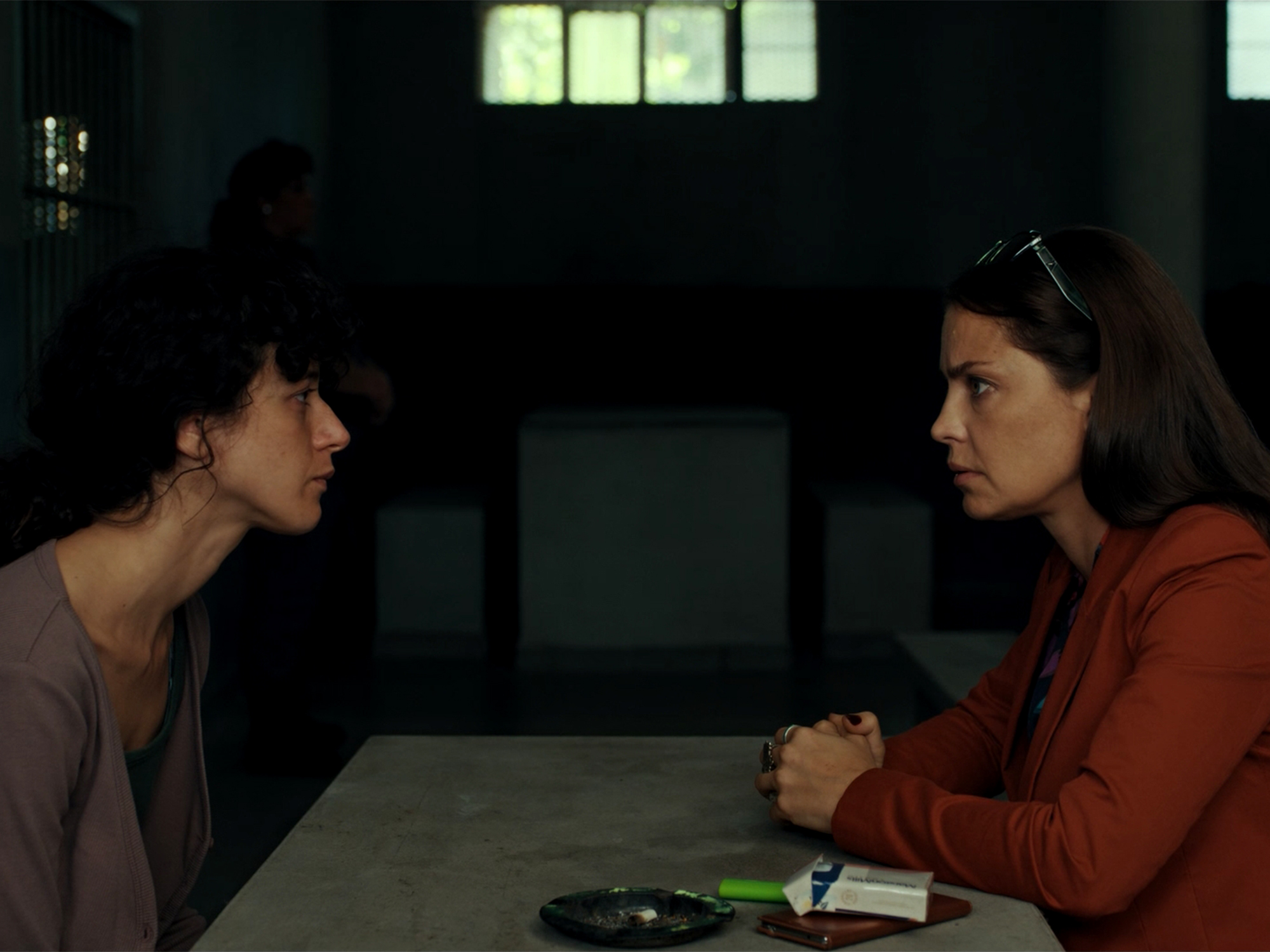 Two women sit across from each other at a table in a dimly lit room with barred windows. One wears grey, the other a red jumper.