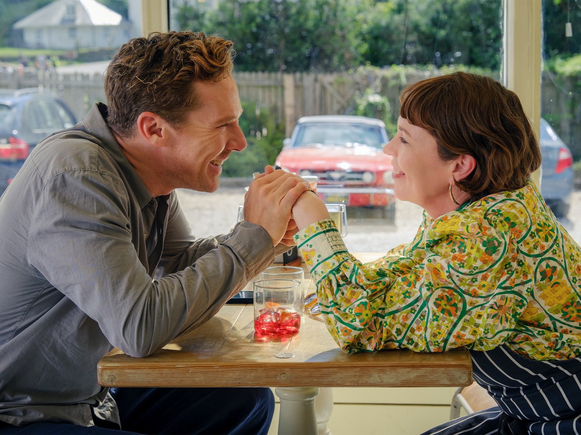 Man in grey suit and boy in yellow patterned shirt sharing a milkshake with two straws at restaurant table by window.