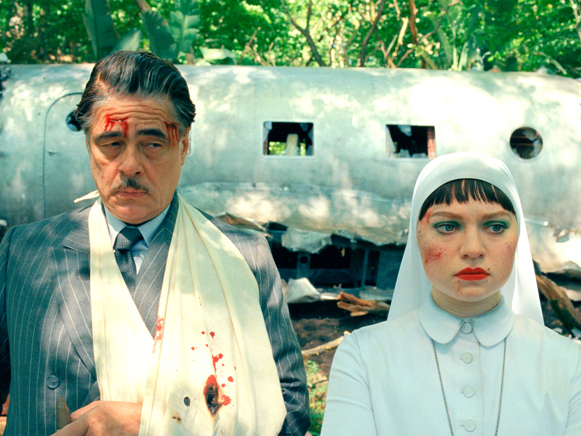 A man in a suit and a woman in a nun's habit stand in front of a damaged trailer in a forest setting.