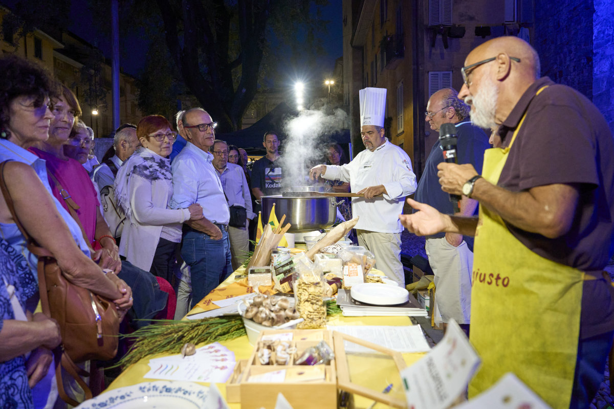 Outdoor evening cooking demonstration with chef in white hat preparing food at table whilst crowd watches under street lighting.