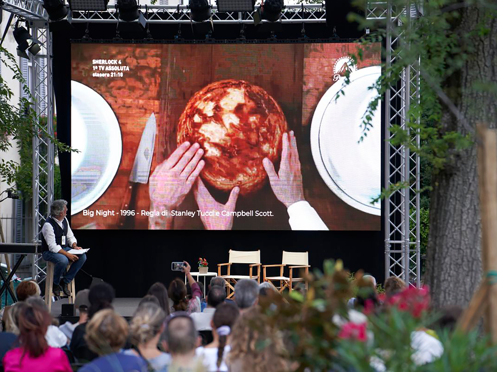 Large outdoor screen displaying red spherical object held by hands, surrounded by white circular shapes. Audience seated below.