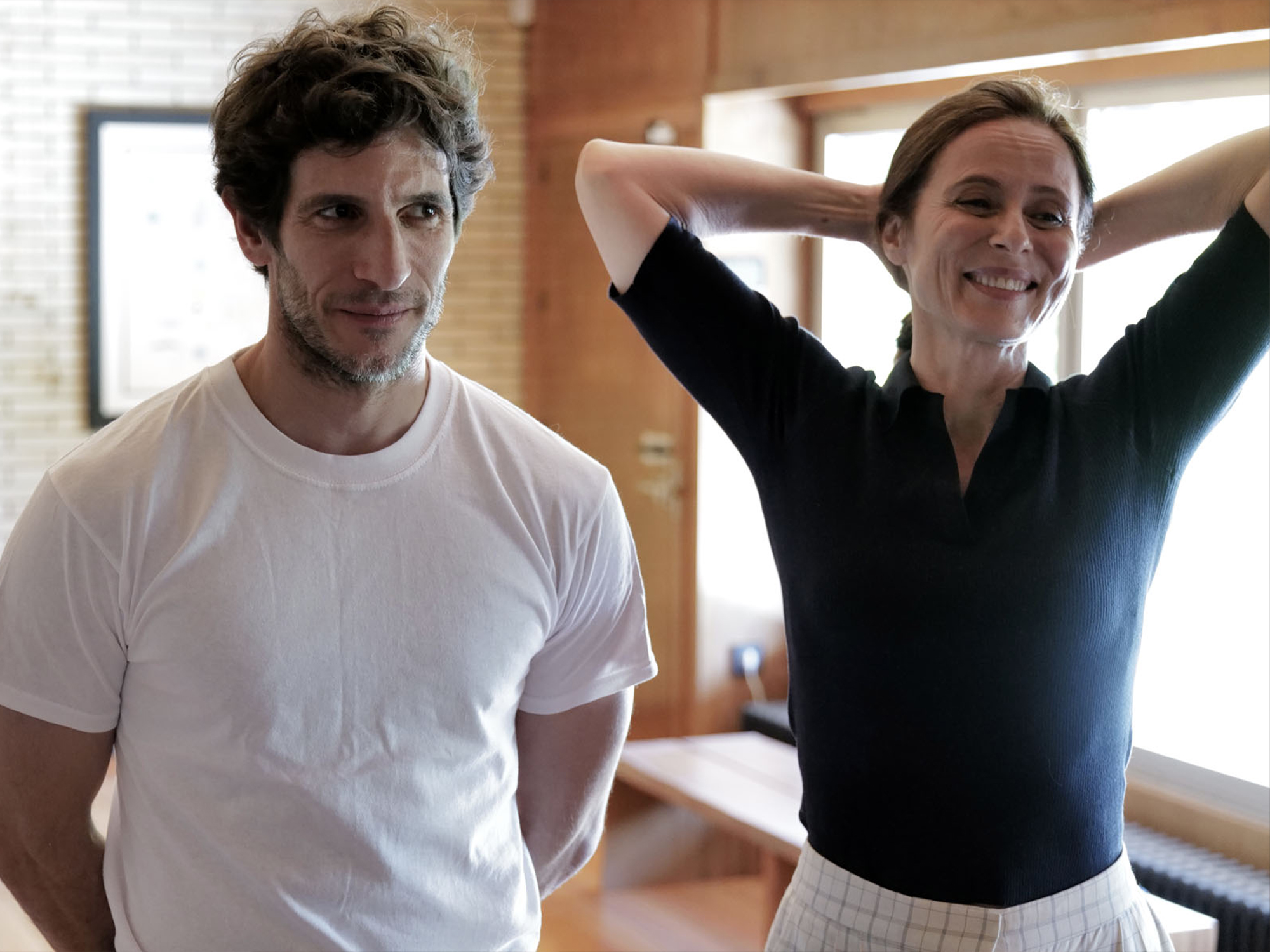Man in white t-shirt and woman in dark top with arms raised, standing in bright interior space with wooden surfaces.