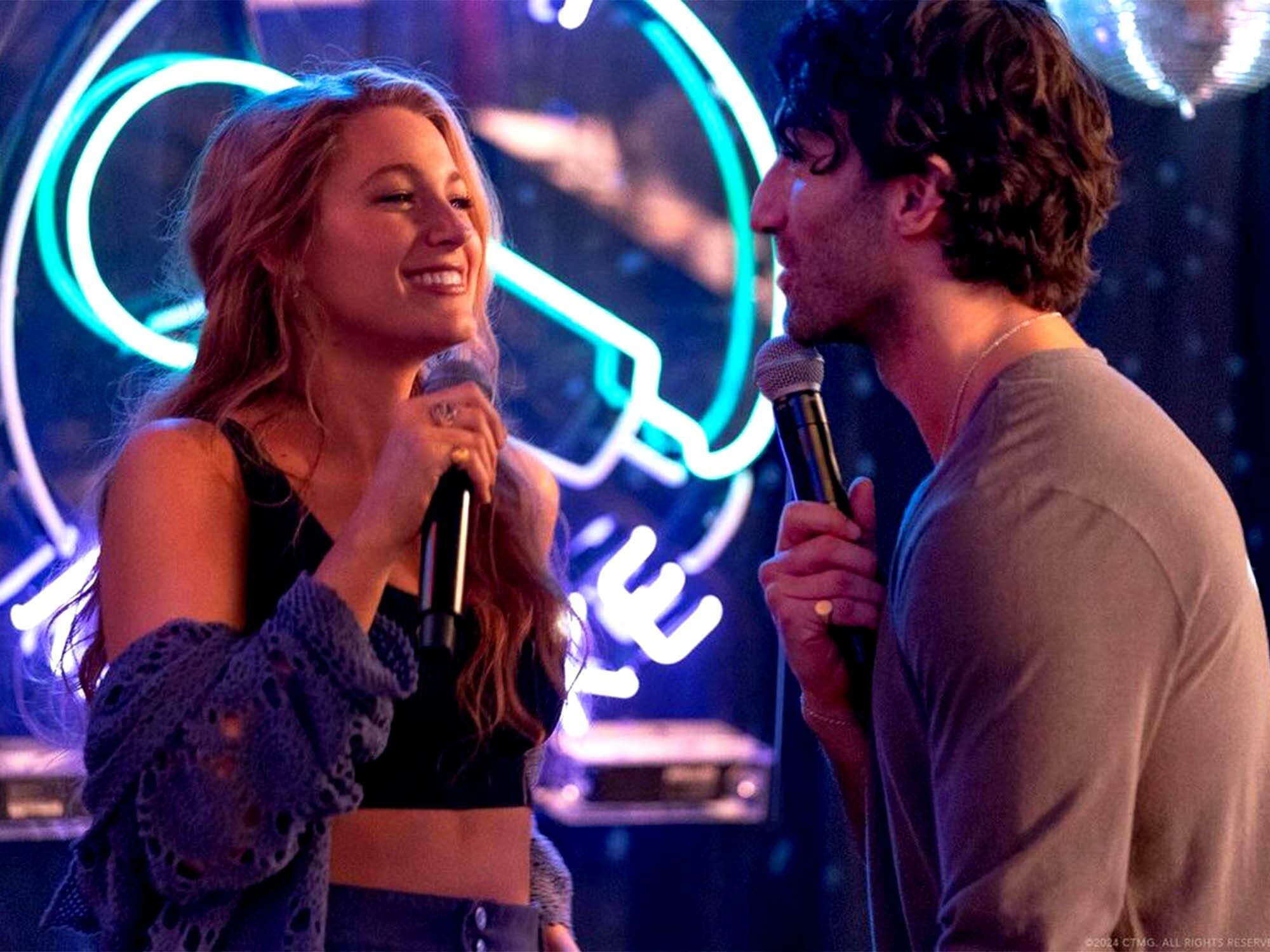 Two people performing on stage, one woman singing into a microphone and one man holding a microphone, against a backdrop of neon lights.