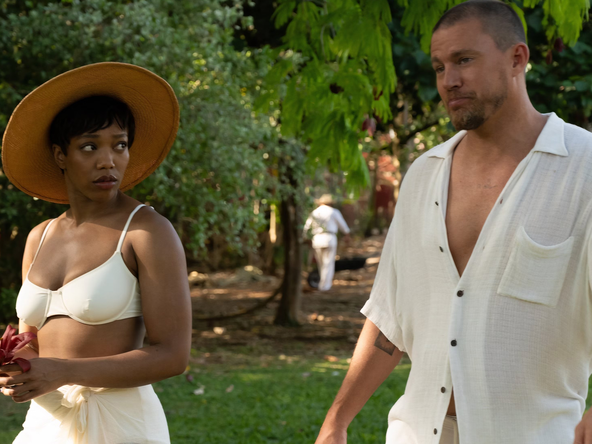 Two people, a woman wearing a sun hat and a man in a white shirt, standing in a lush, green garden.